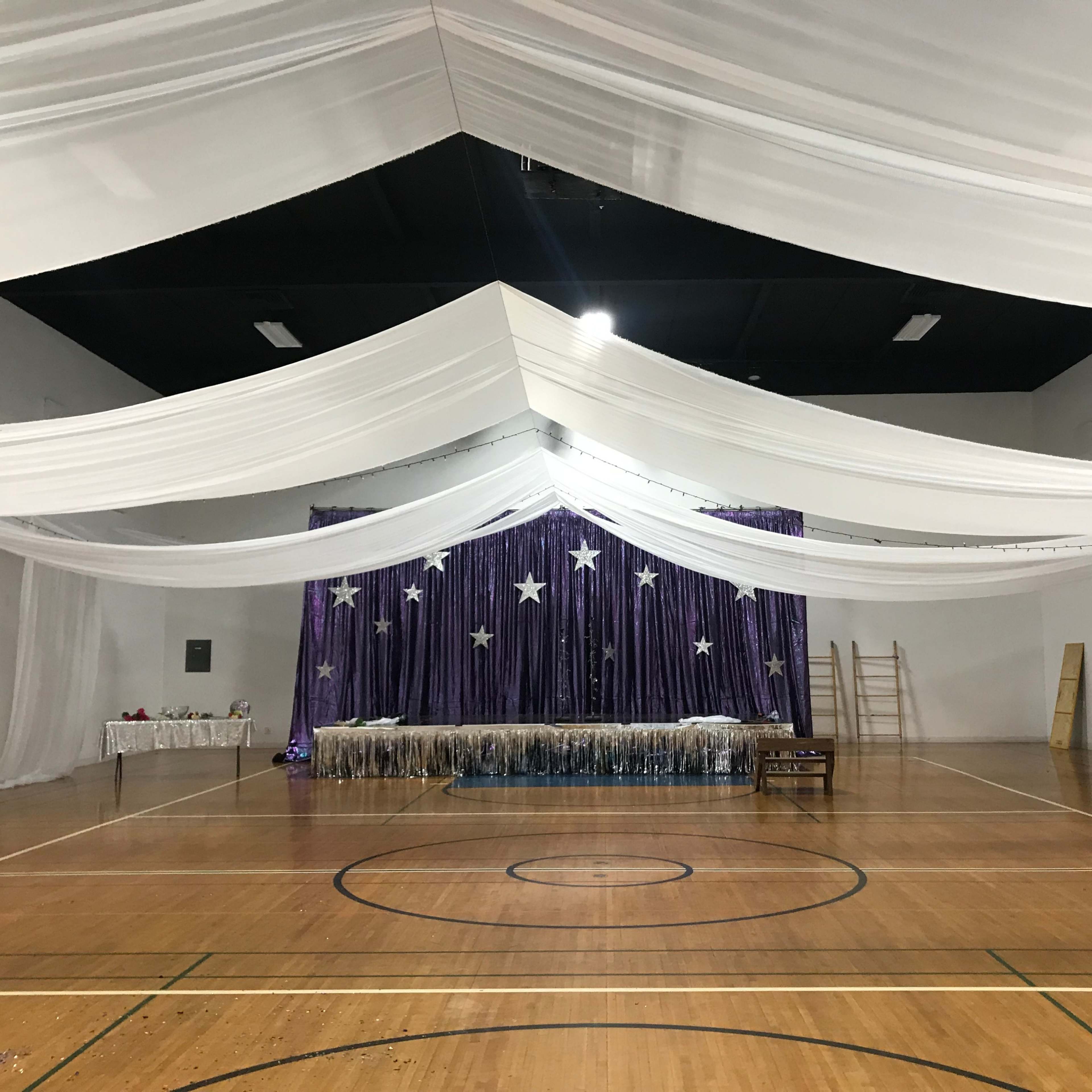A decorated indoor space with white drapery overhead, a purple backdrop with stars, and a table in front, all set up for an event.
