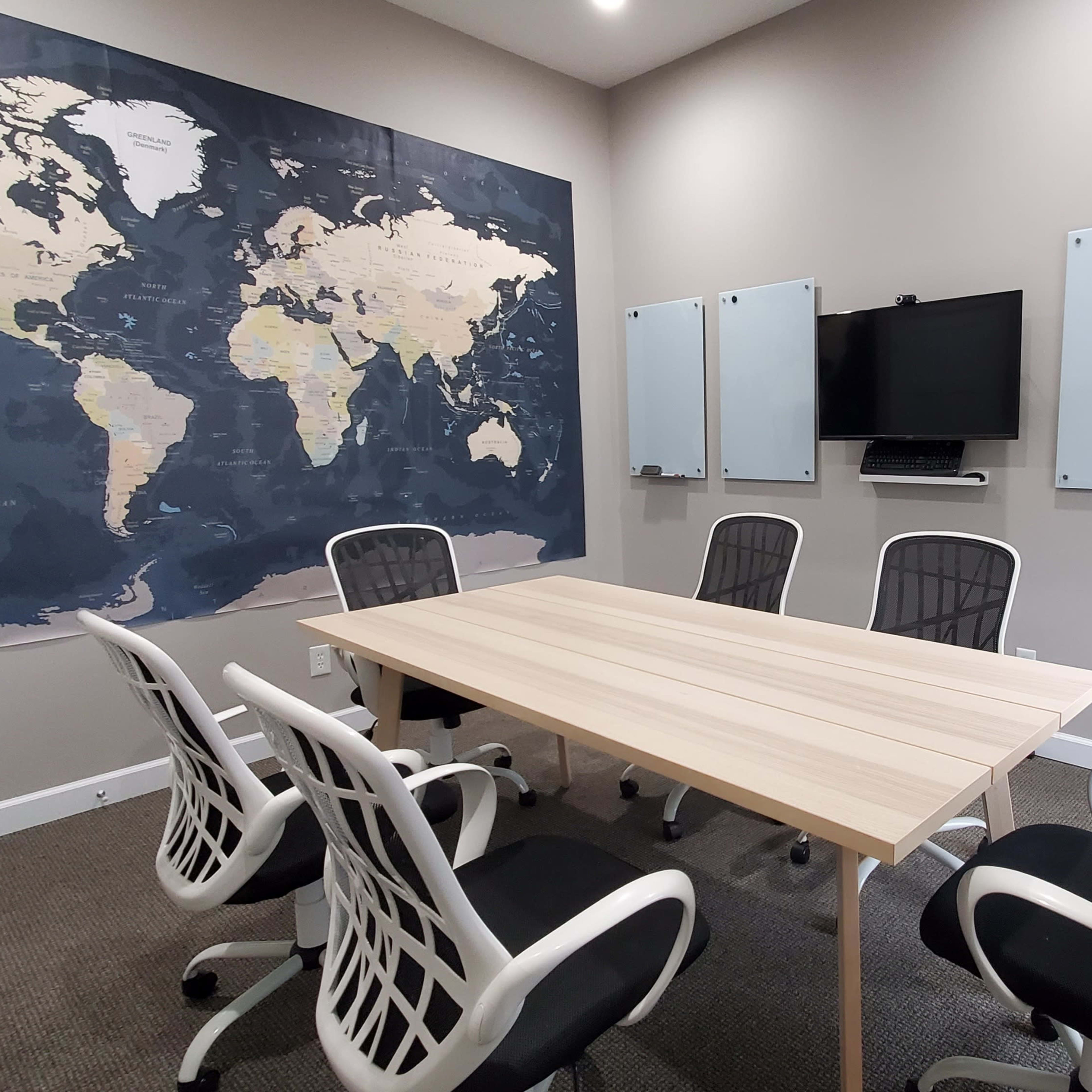 A conference room features a large wooden table surrounded by five chairs, with a world map on the wall and several whiteboards on either side.