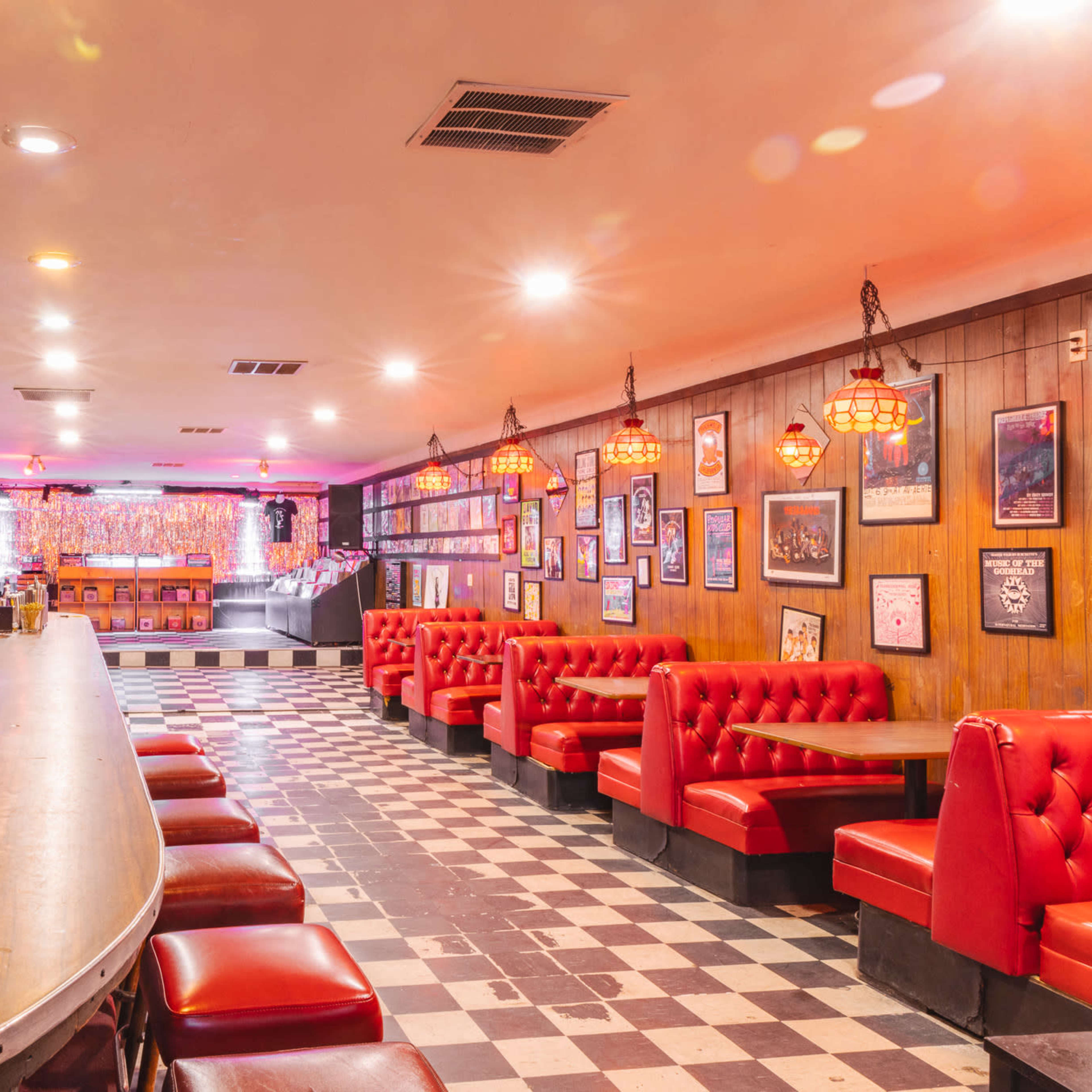 A vintage-themed bar with a long wooden counter, red booths, and checkered flooring, featuring dim lighting and various decorations on the walls.