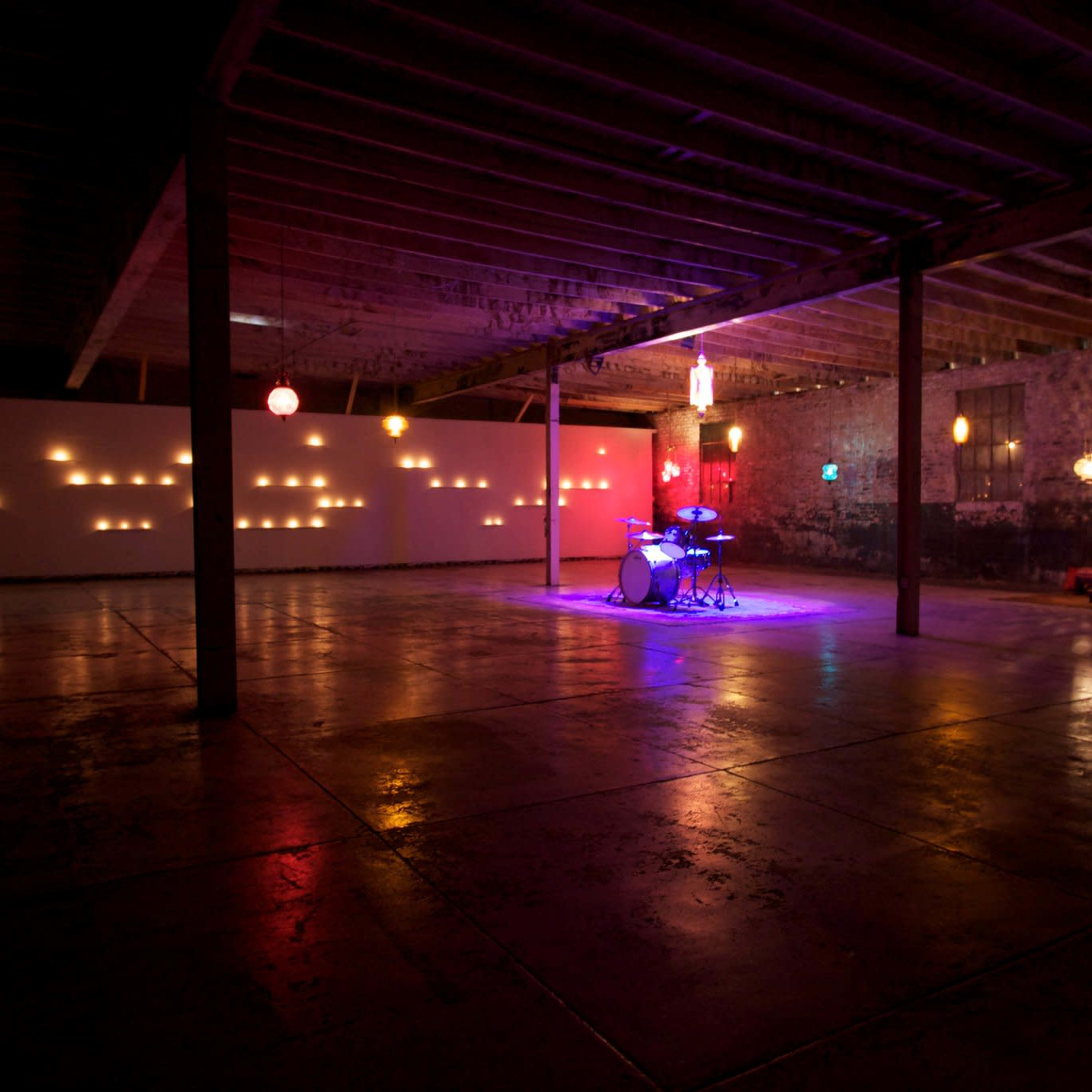 A spacious, dimly lit warehouse features a drum set illuminated by colored lights, with wall projections and hanging lamps casting soft glows around the room.