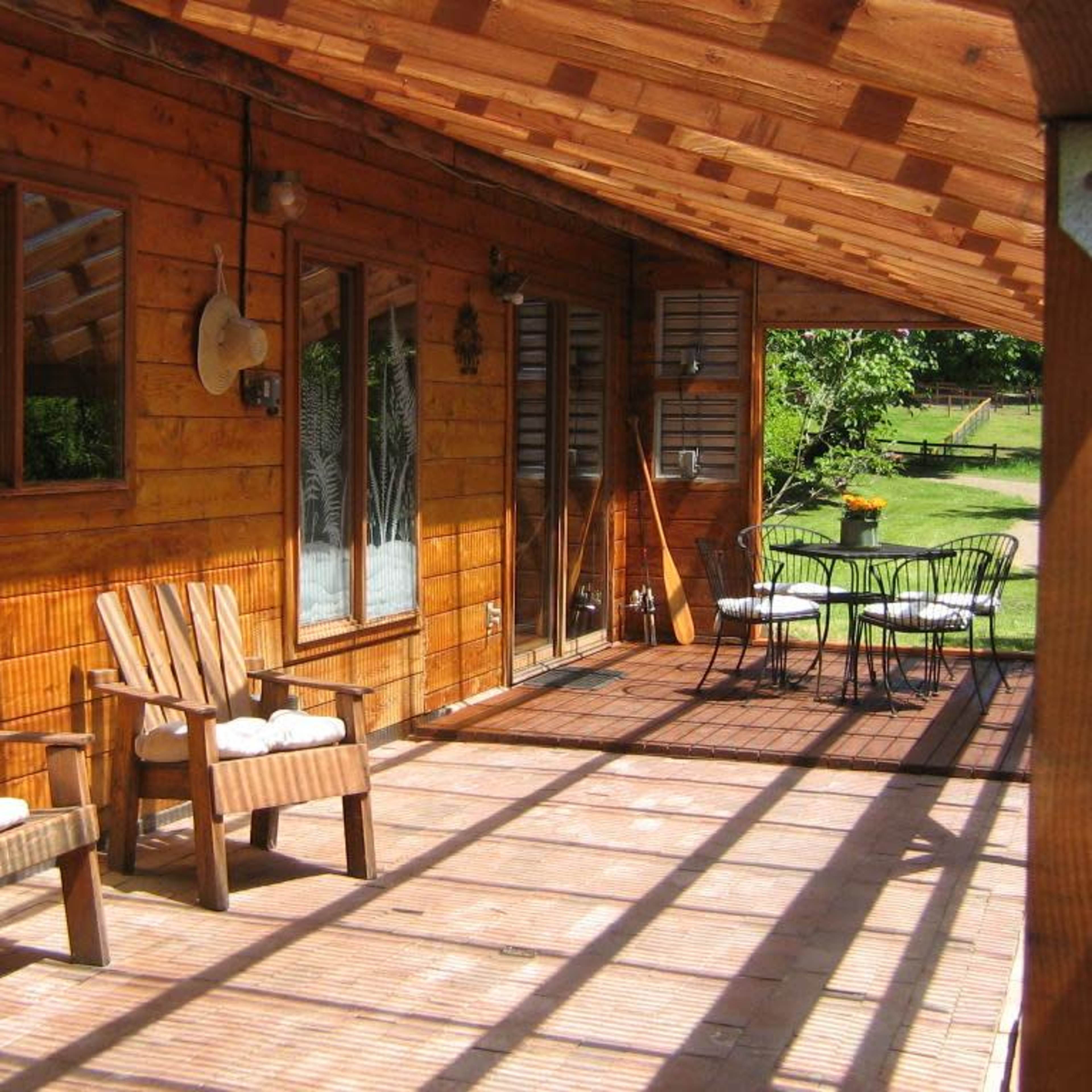 A wooden porch features two chairs and a round table set against a backdrop of grassy outdoor space.