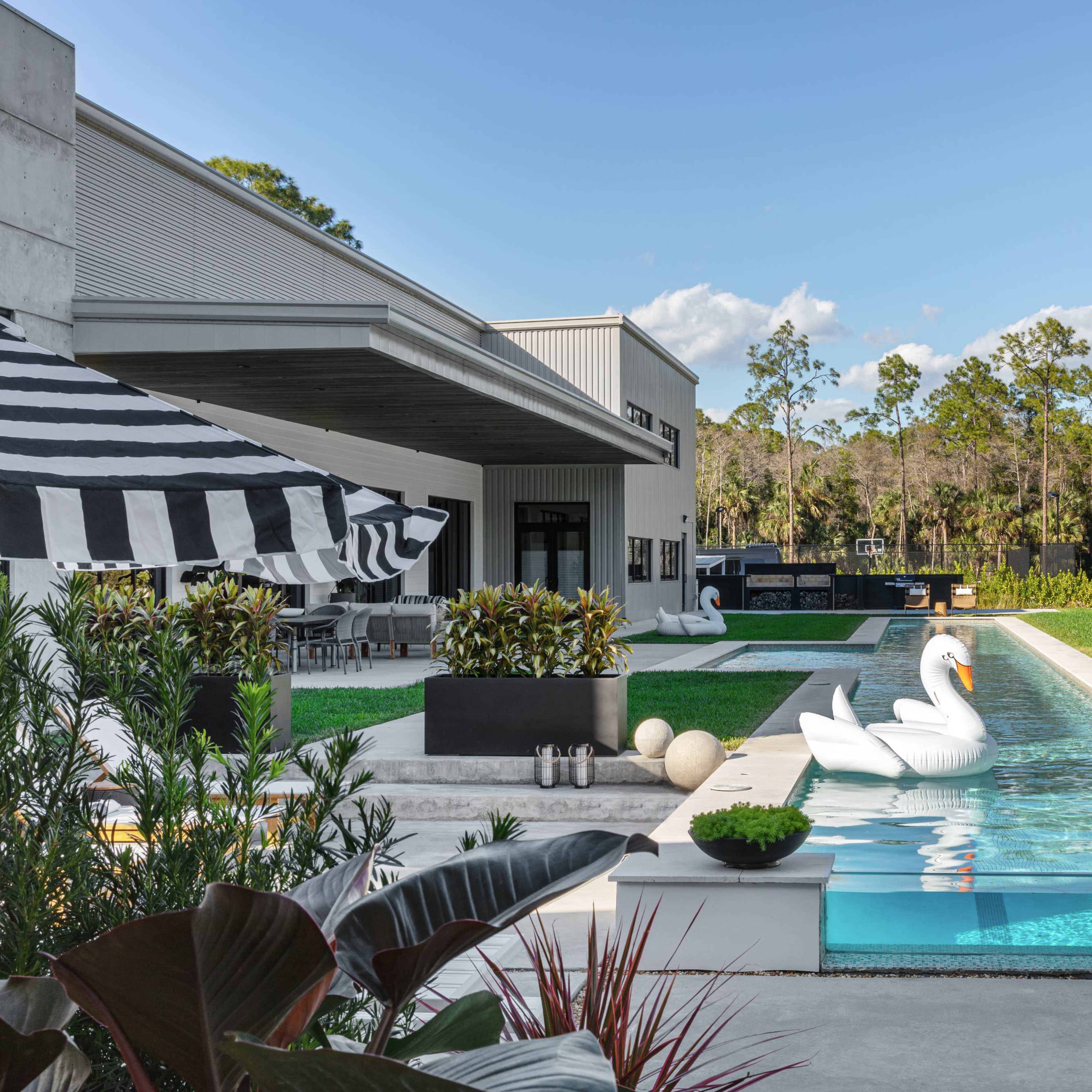 A modern outdoor space featuring a swimming pool with a swan float, surrounded by a landscaped lawn, lounge areas, and a contemporary structure.