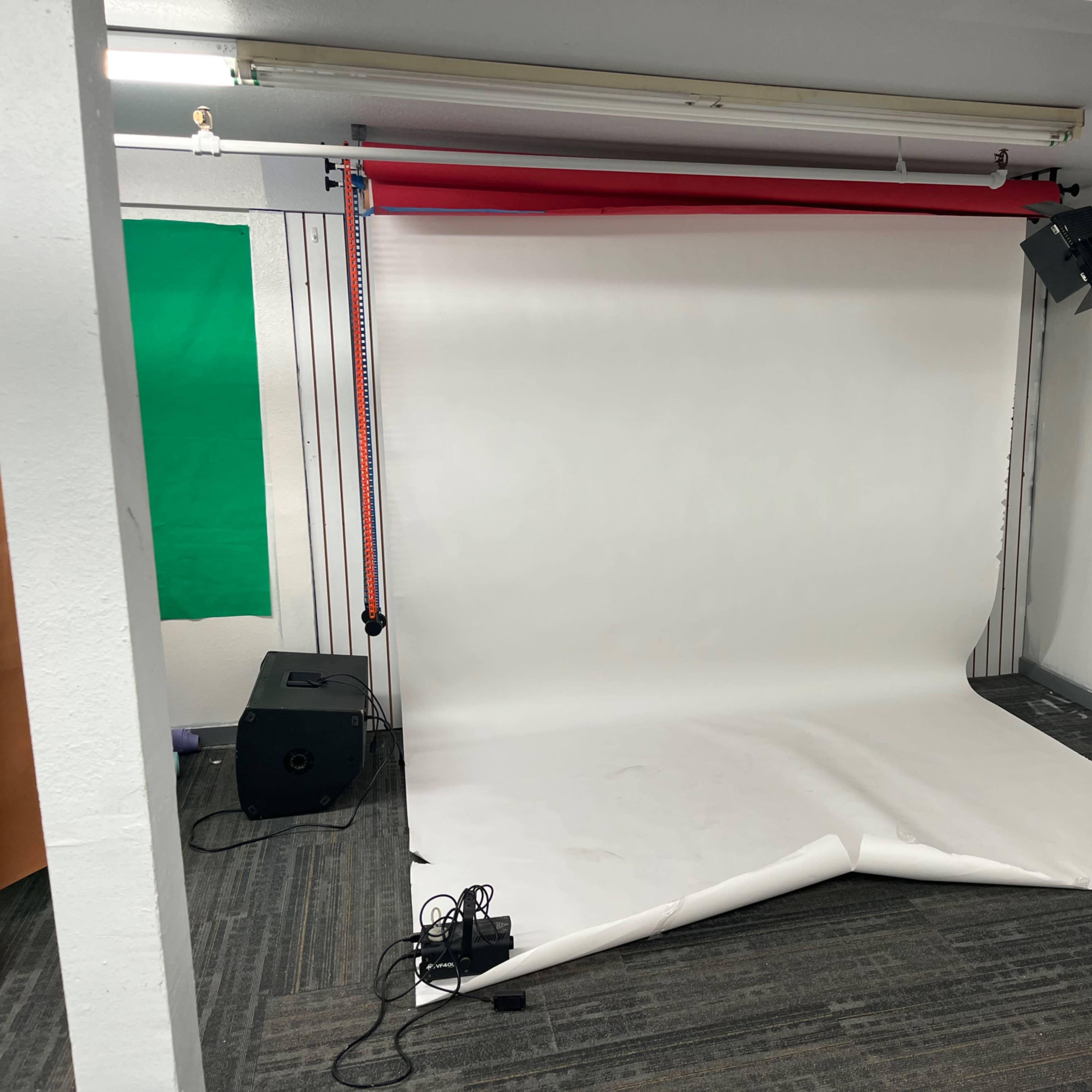 A photography setup featuring a white backdrop, a light stand with a light source, and equipment on the floor in a room with gray carpet.