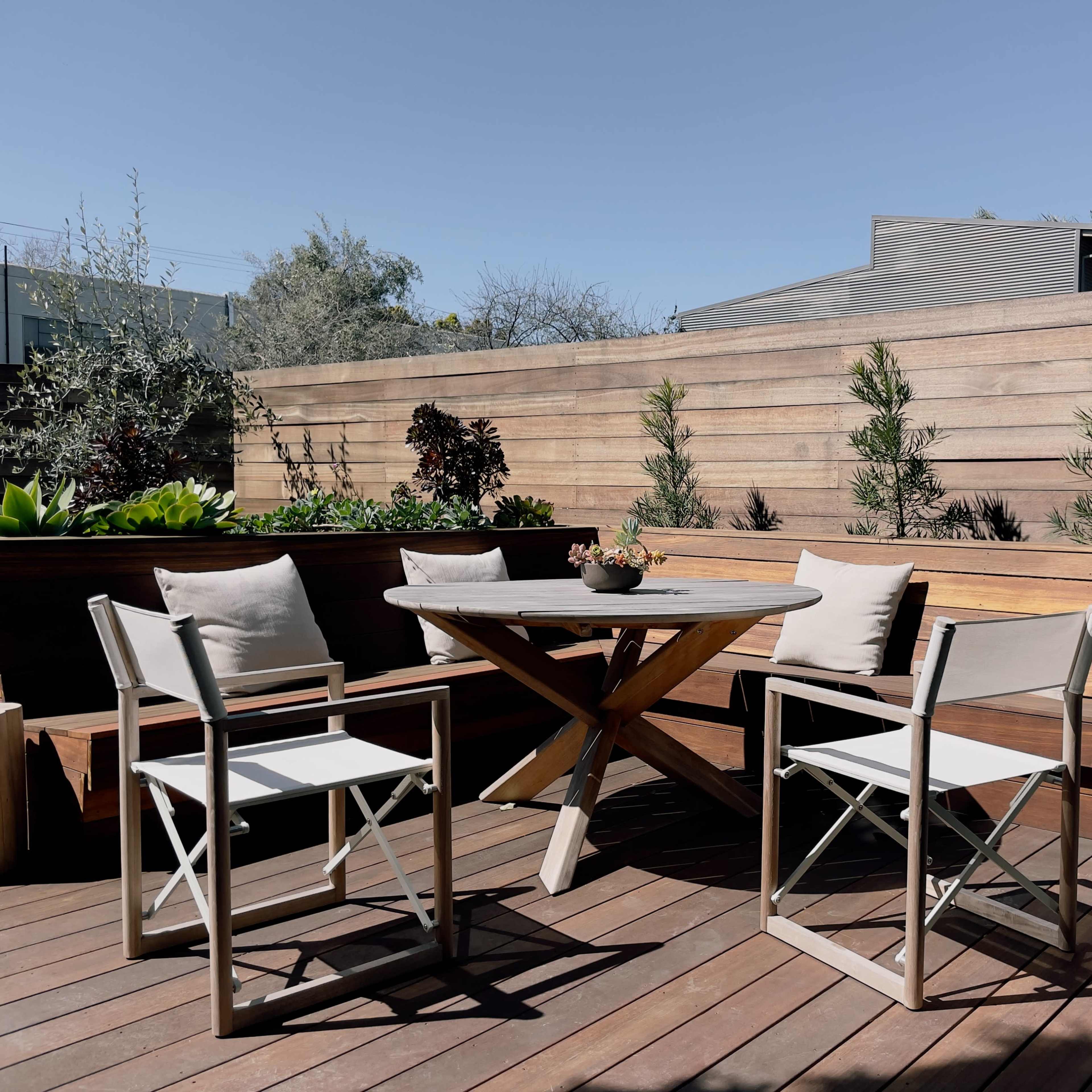 A wooden deck features a round table surrounded by four folding chairs, with planters and greenery in the background.