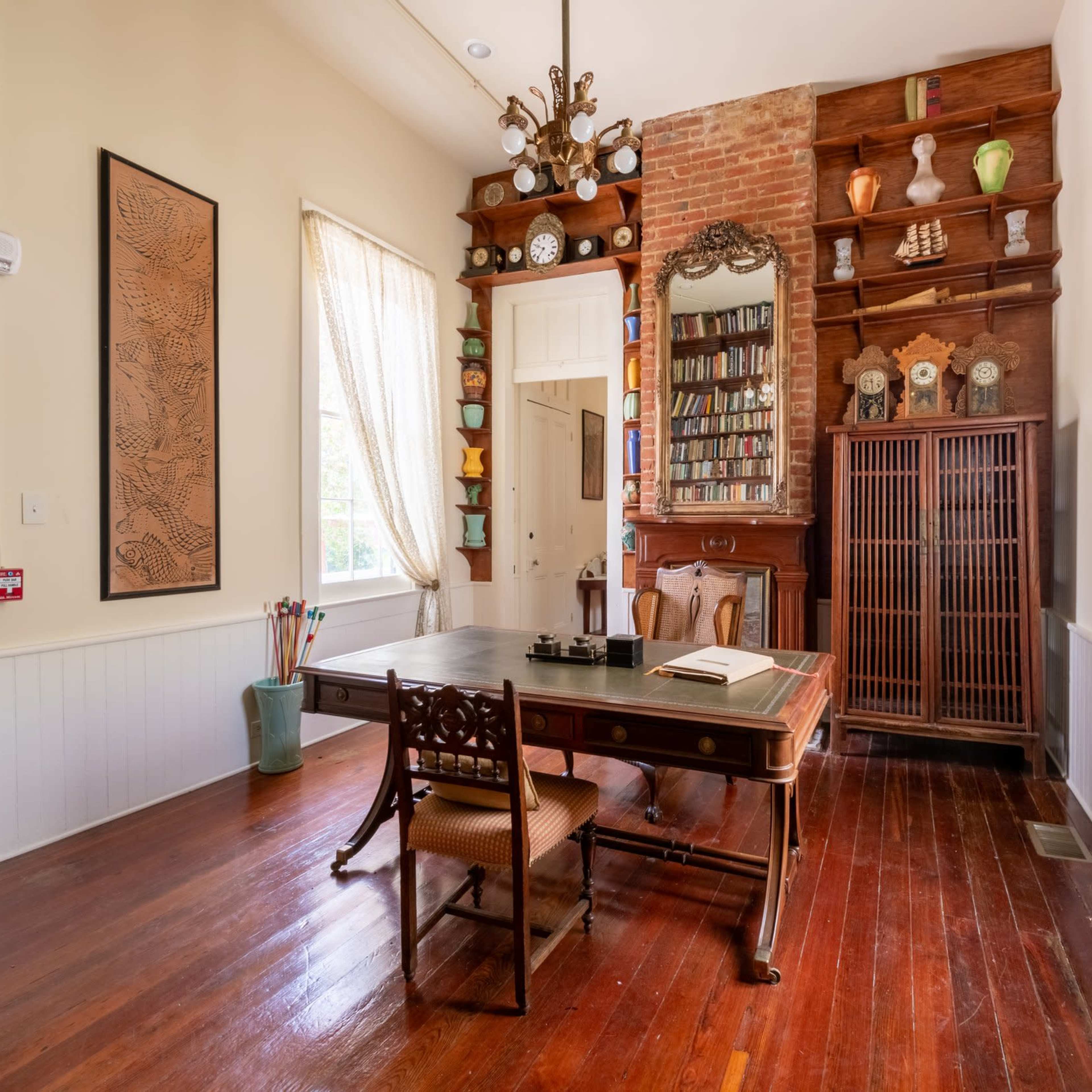 A wooden table with chairs in a room featuring a bookshelf, decorative shelves, and large windows with light curtains.