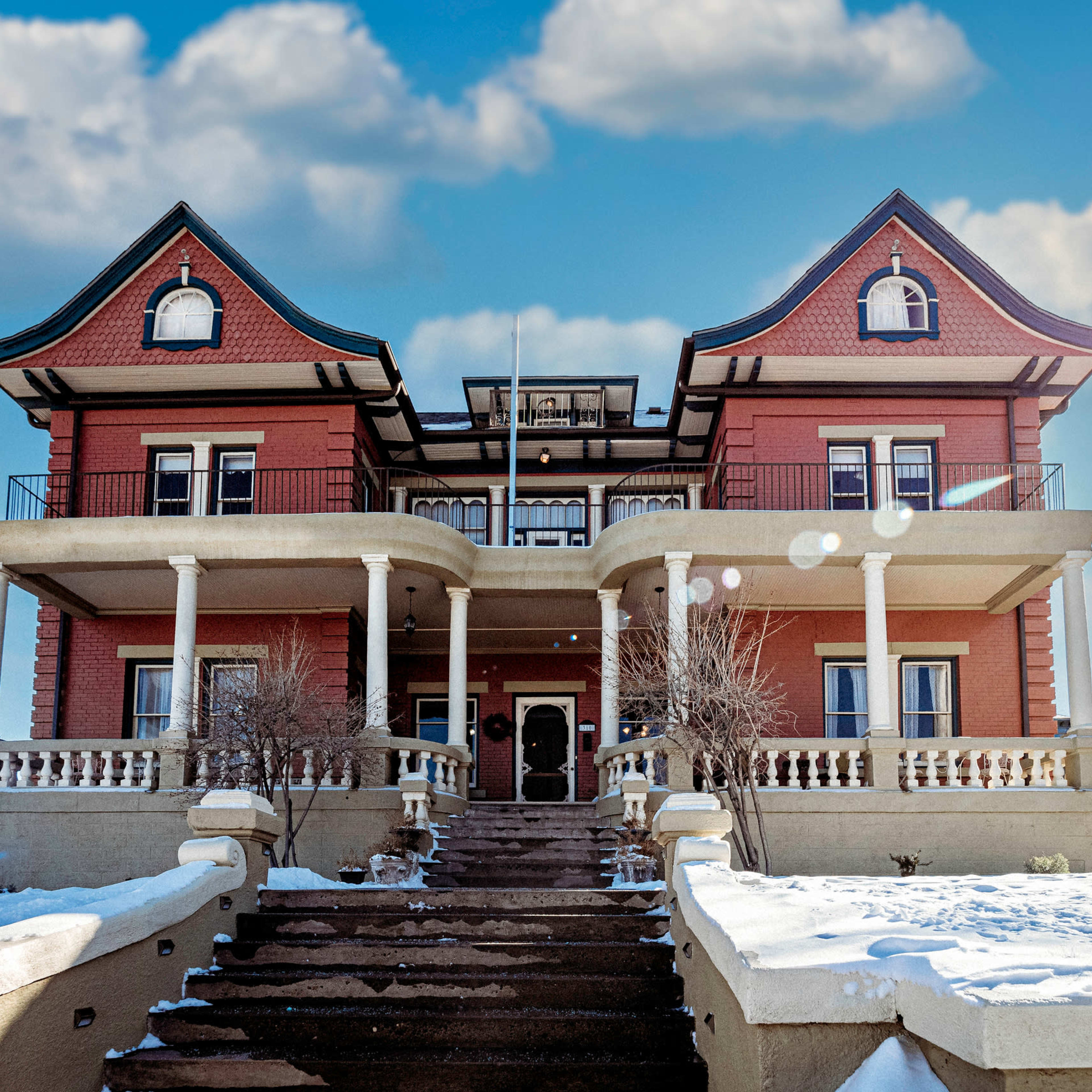 A large, Victorian-style house with a red exterior, multiple balconies, and a staircase leading to the entrance, set against a partly cloudy sky.