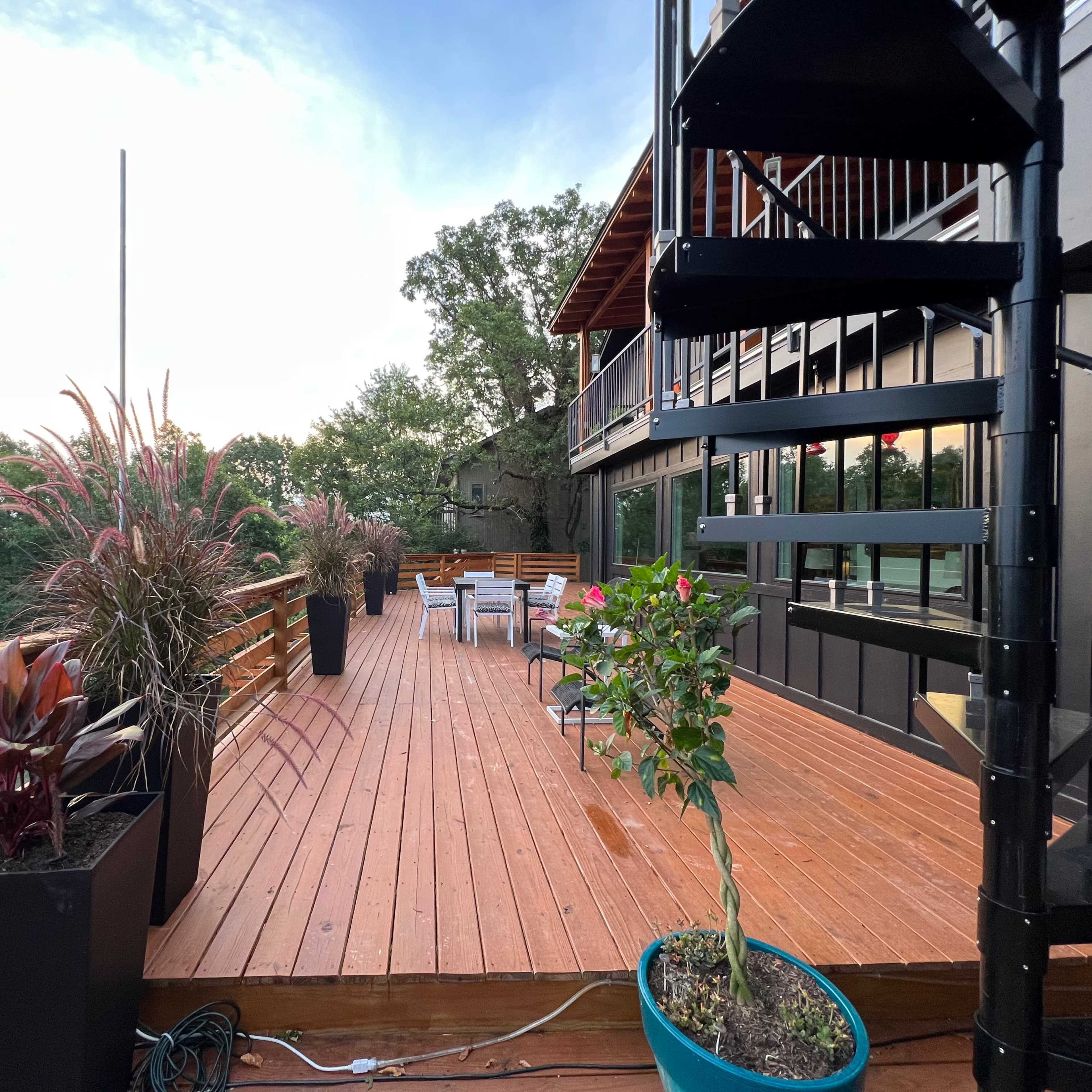 A wooden deck with potted plants and a spiral staircase, overlooking a forested landscape under a cloudy sky.