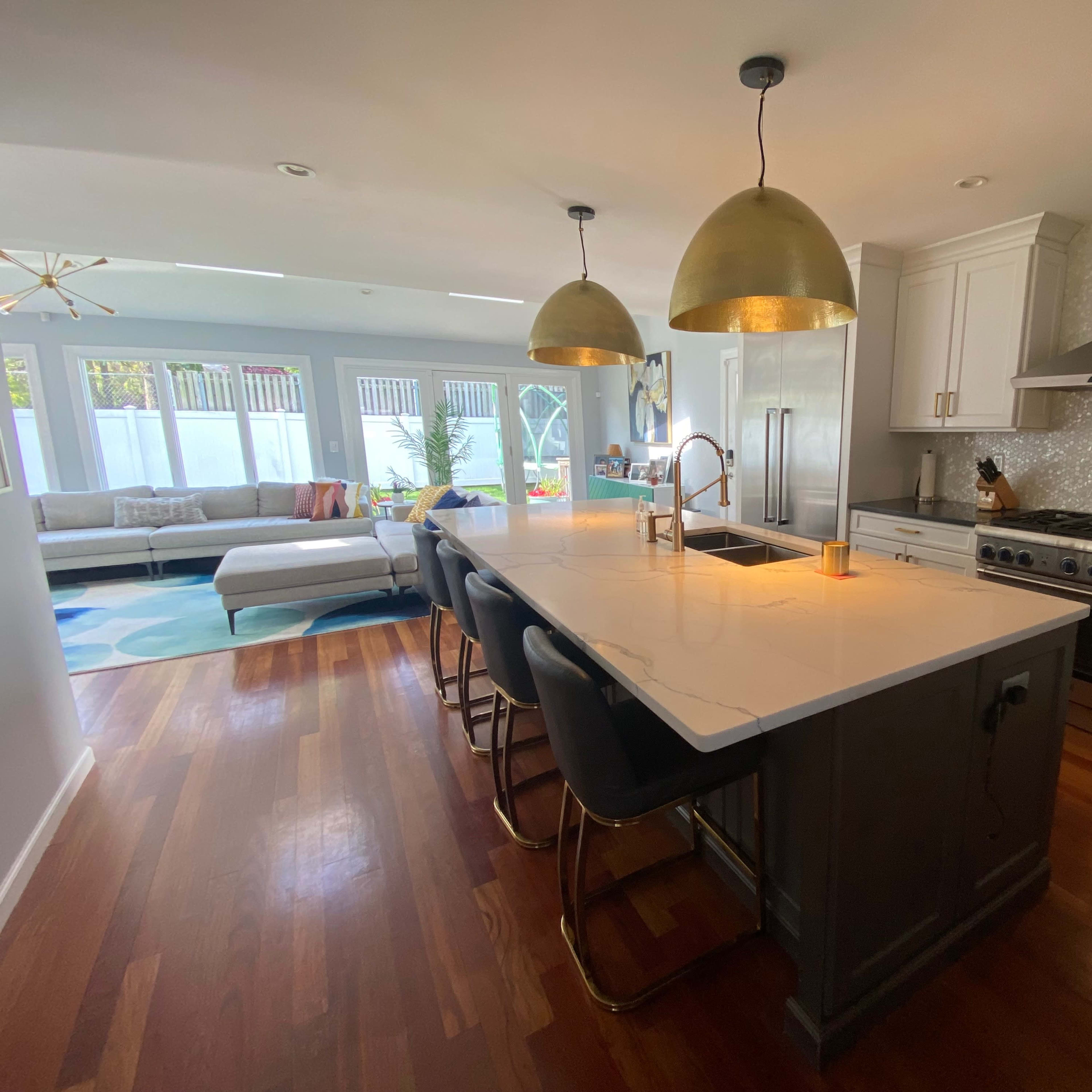 A modern kitchen with a large island featuring seating, pendant lights, and stainless steel appliances, leading into a living area with a sectional sofa.