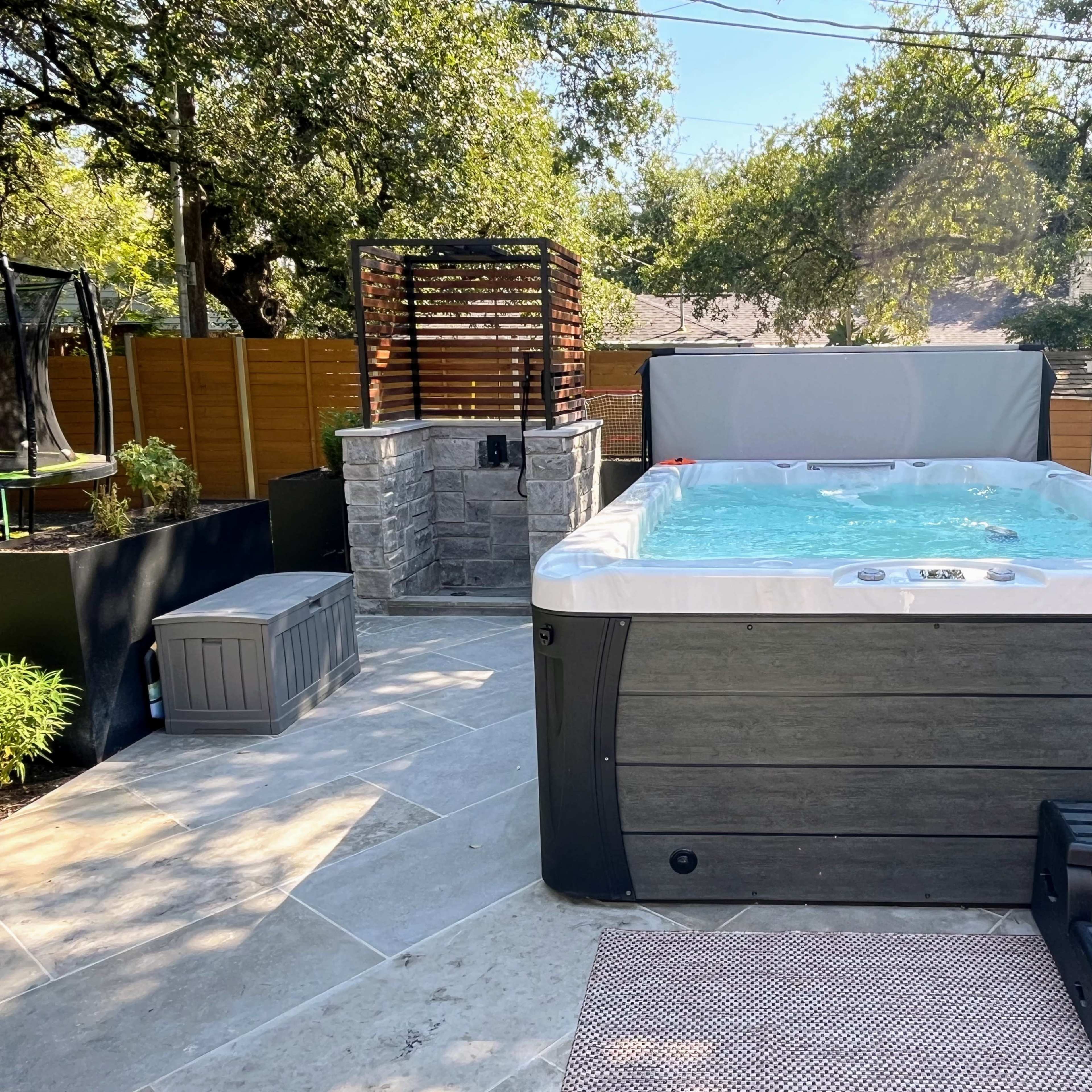 A backyard scene featuring a hot tub surrounded by stone and wood elements, with a small seating area and greenery in the background.