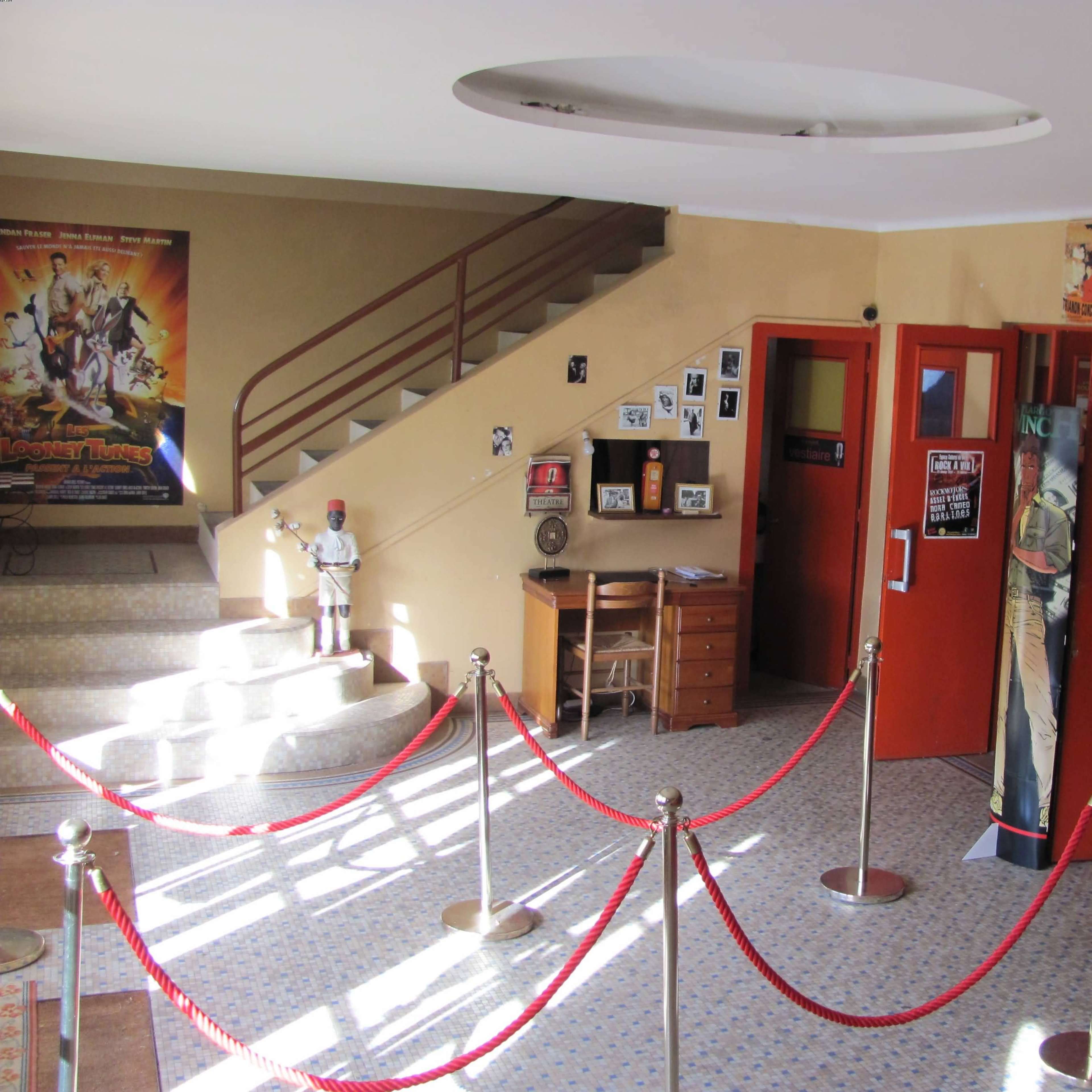 An interior view of a vintage movie theater lobby with a staircase, red velvet ropes, and posters on the walls.