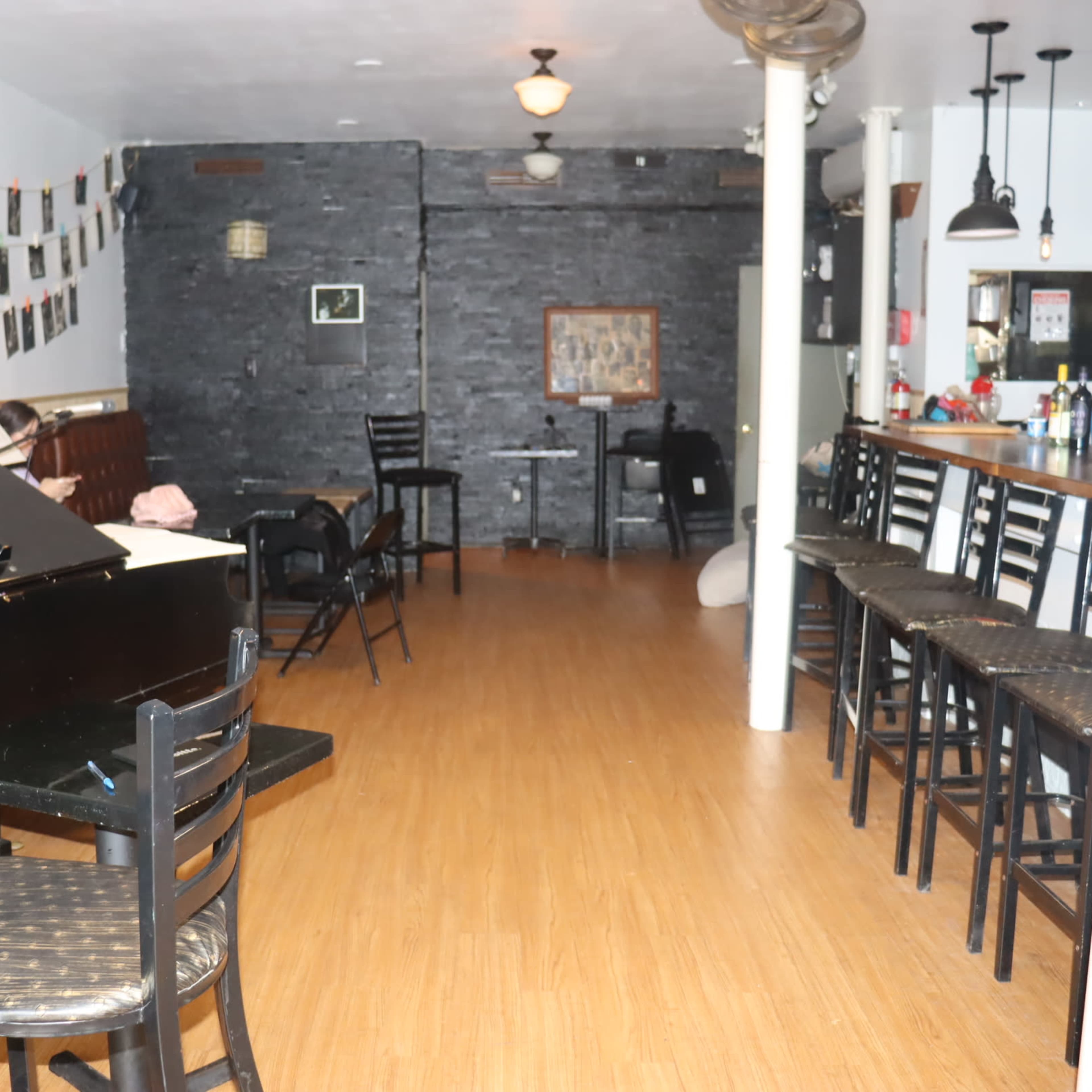The interior of a music venue features a grand piano in the foreground, rows of black chairs, and a bar against the back wall.