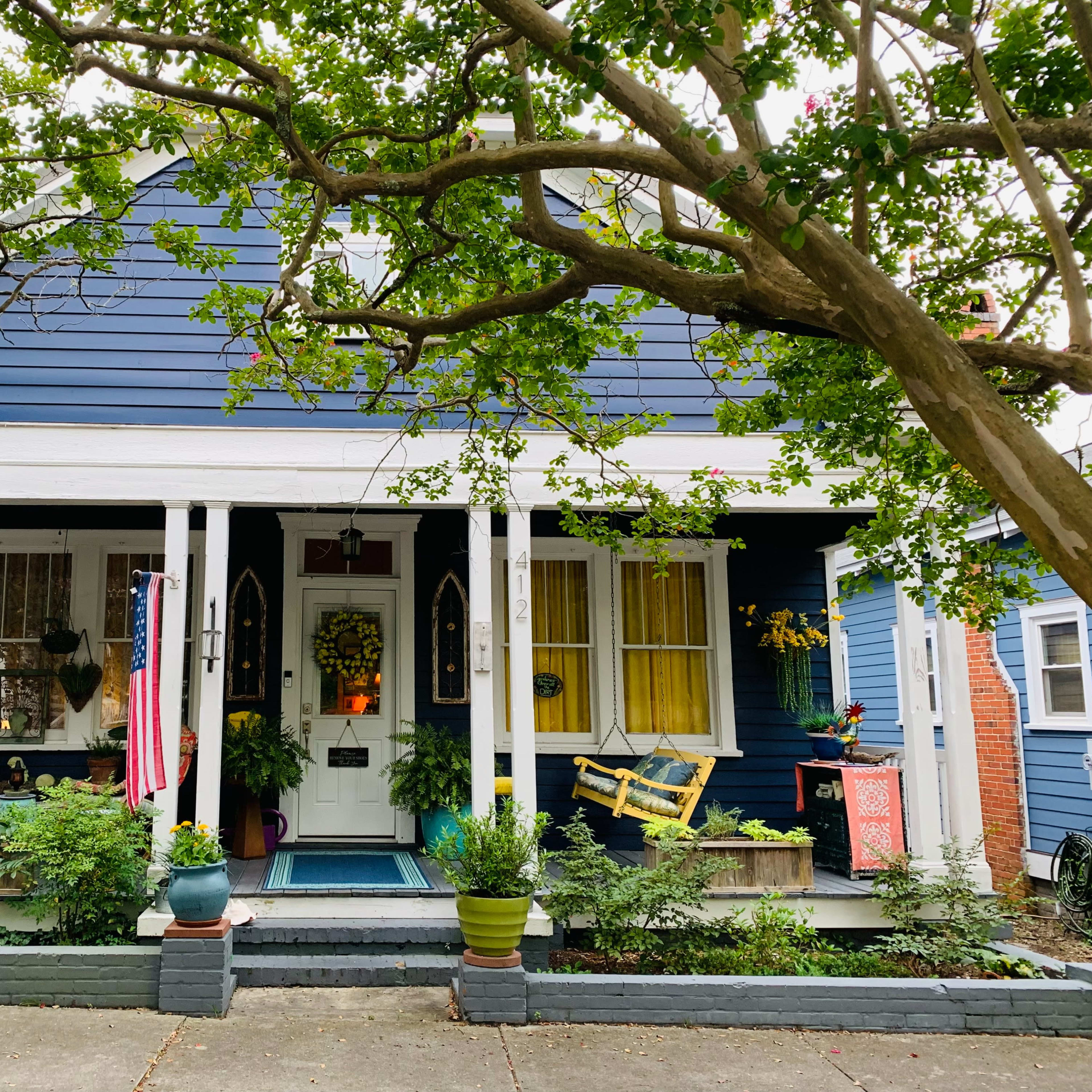 A blue house with white columns and porch, adorned with plants and an American flag, sits beneath a large tree.