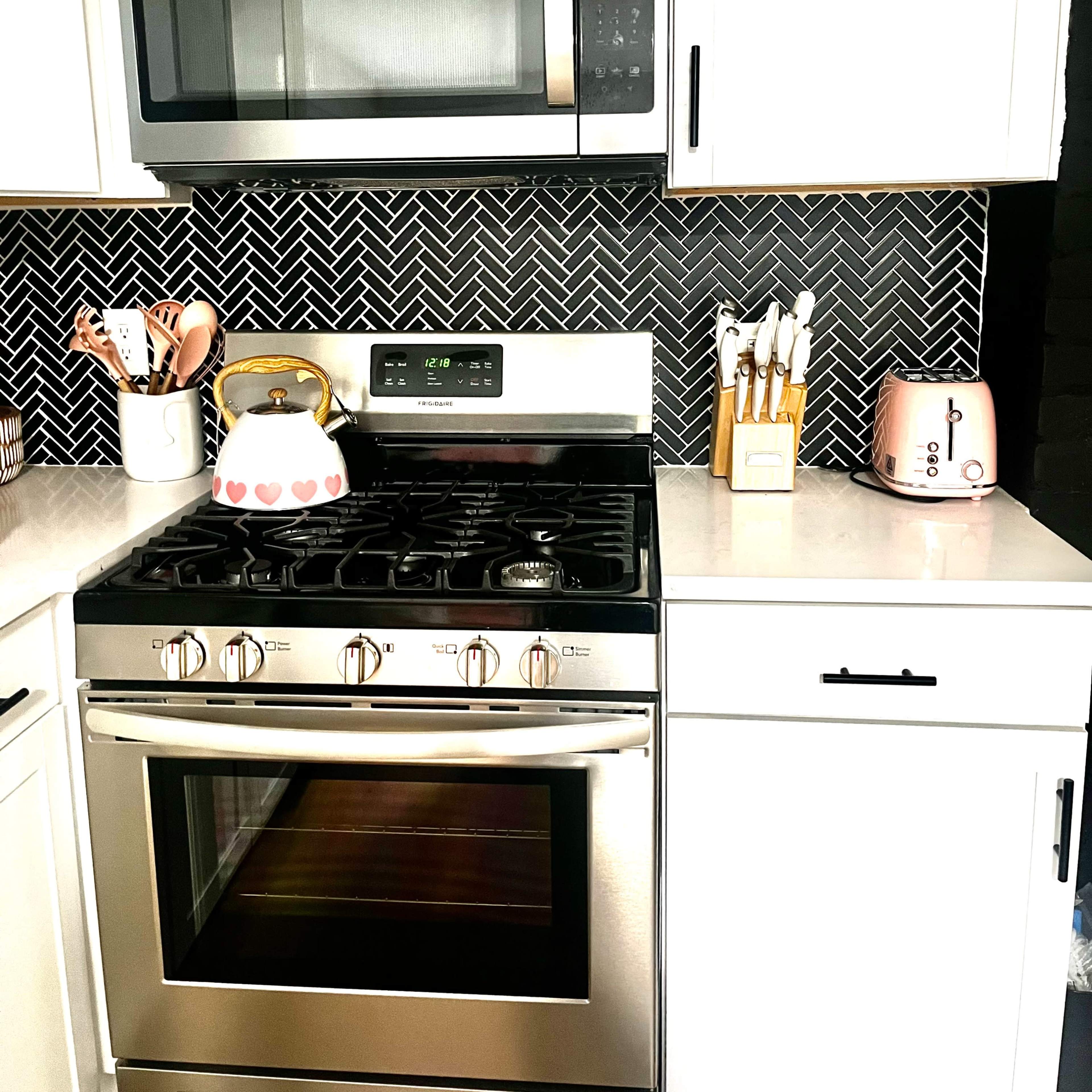 A modern kitchen featuring stainless steel appliances, including a gas stove and microwave, against a patterned backsplash with white cabinets and a pink toaster.