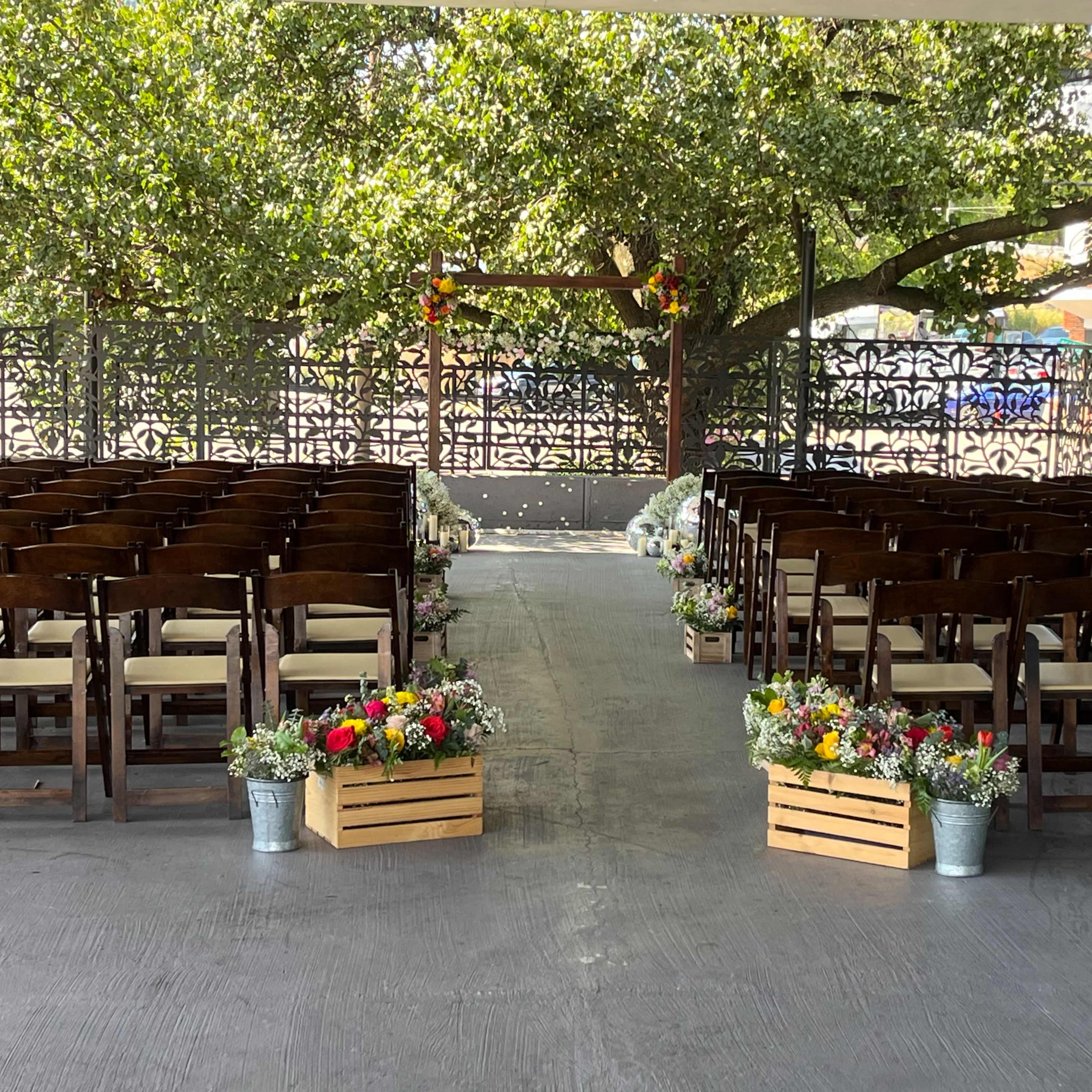 A wedding ceremony setup with rows of wooden chairs facing an arched structure decorated with flowers, framed by greenery.
