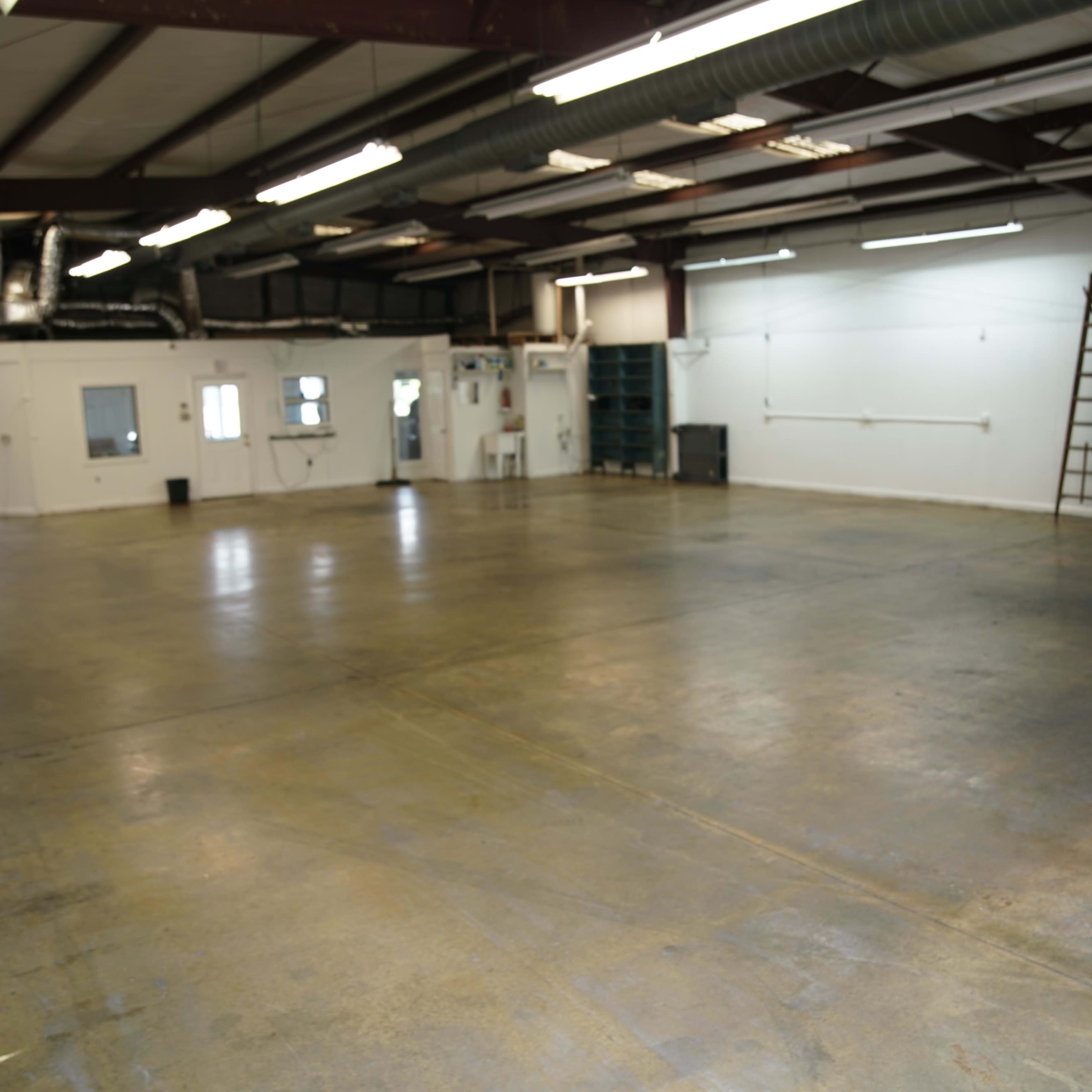 A spacious industrial room with a concrete floor, several windows, and a ladder against one wall.