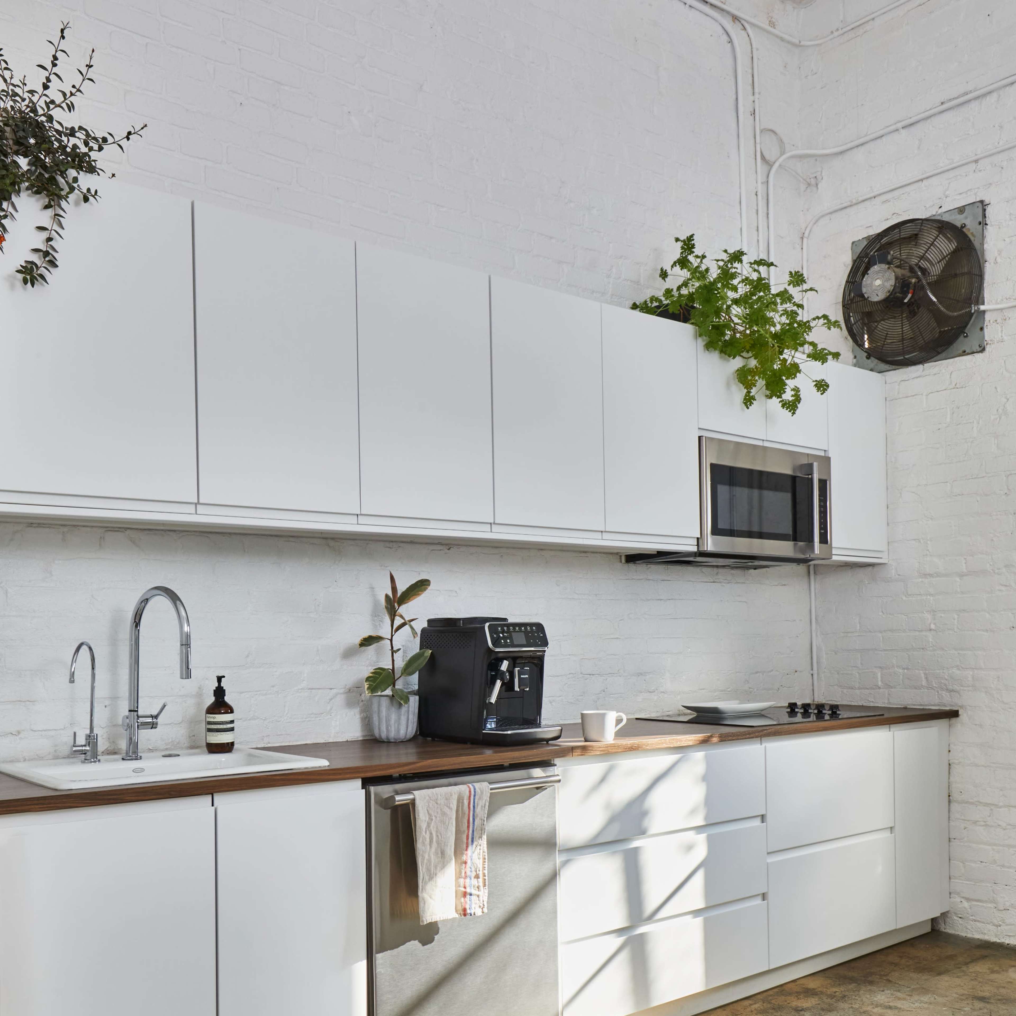 A modern kitchen with white cabinetry, a stainless steel refrigerator, a coffee machine, and plants, illuminated by natural light from a nearby window.