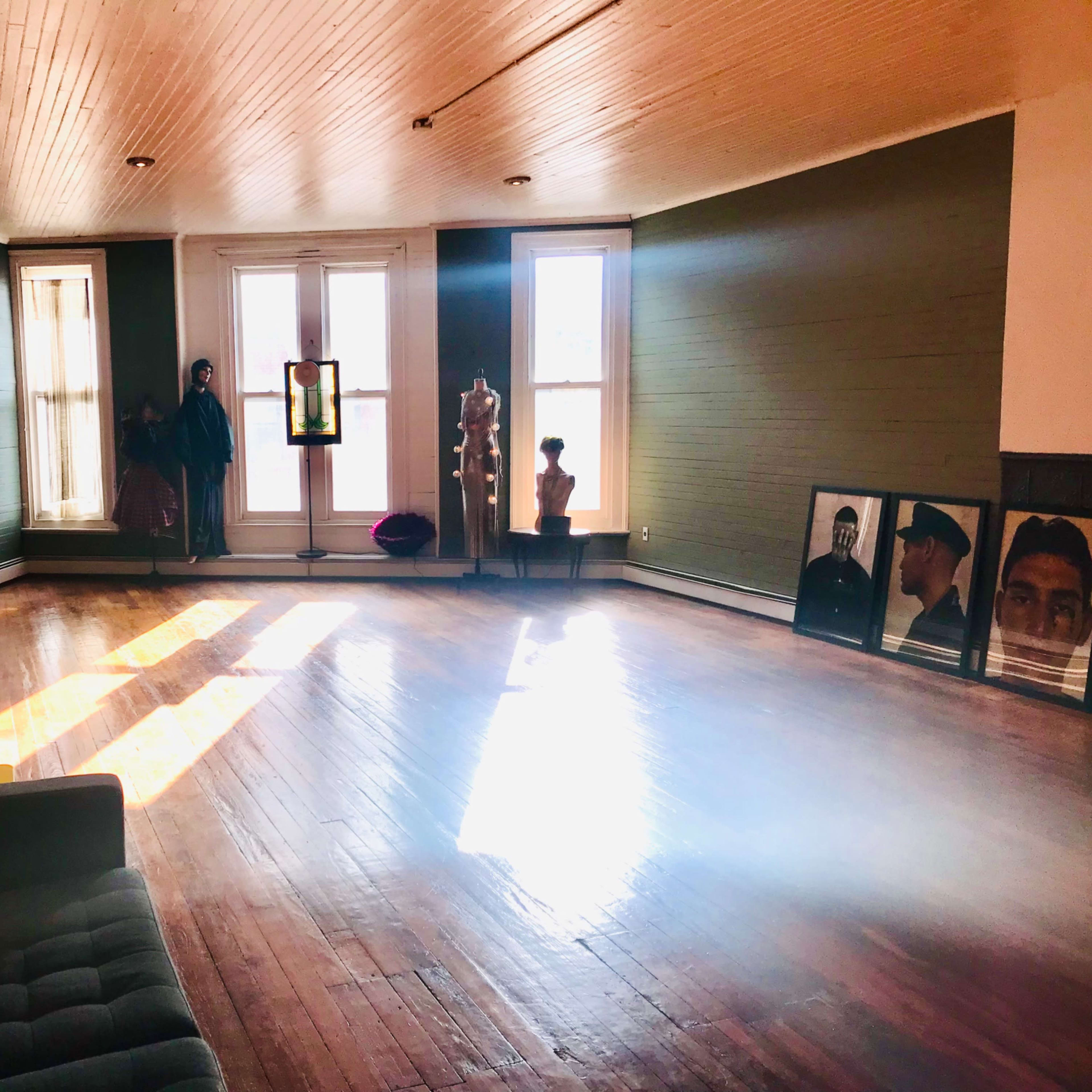 A spacious room with wooden floors and large windows, featuring a sofa, a mannequin, and framed photographs on the floor.