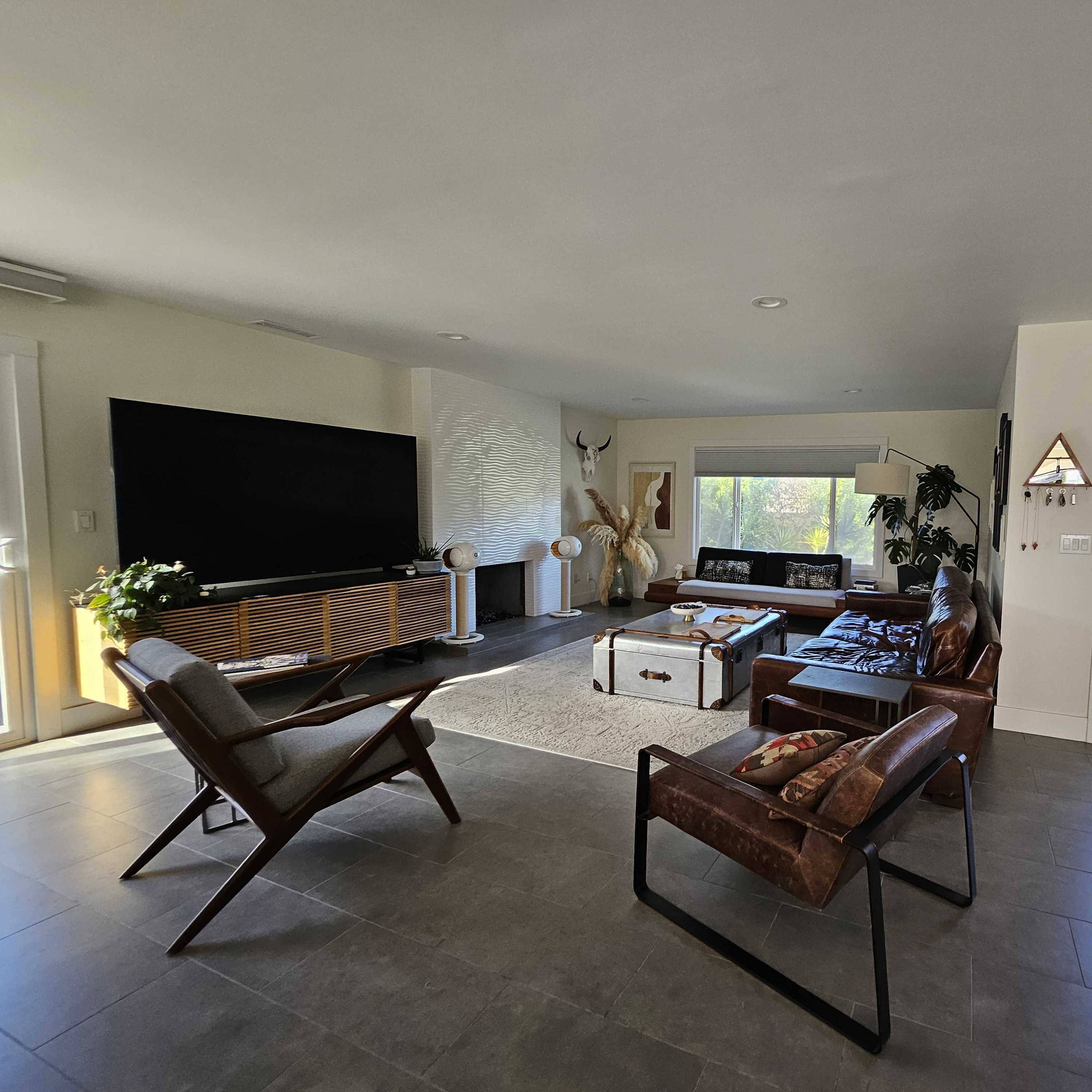 A modern living room featuring a large television, a coffee table, two armchairs, and a door leading outside.