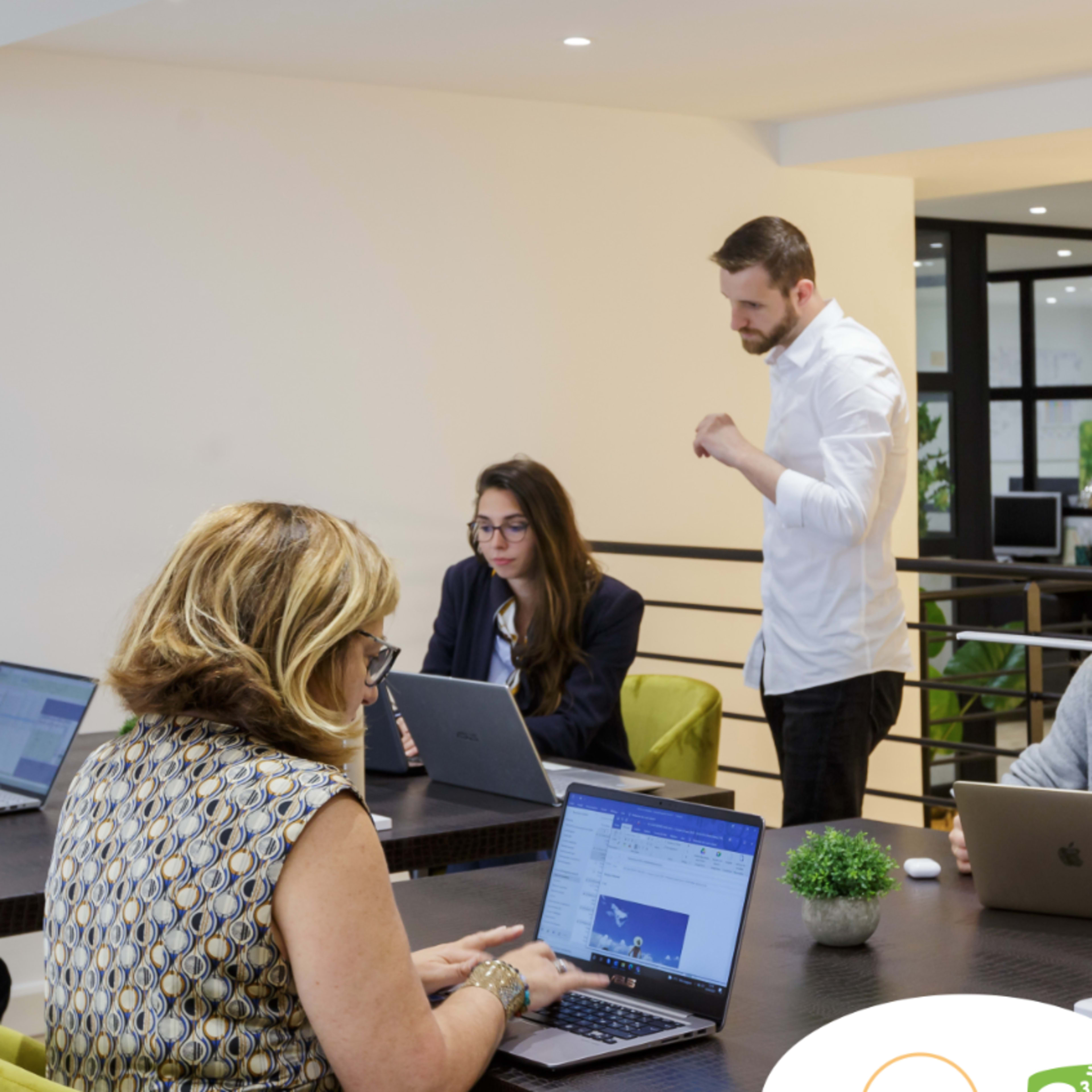 A group of five people is collaborating in a modern office setting, working at laptops while discussing a project.