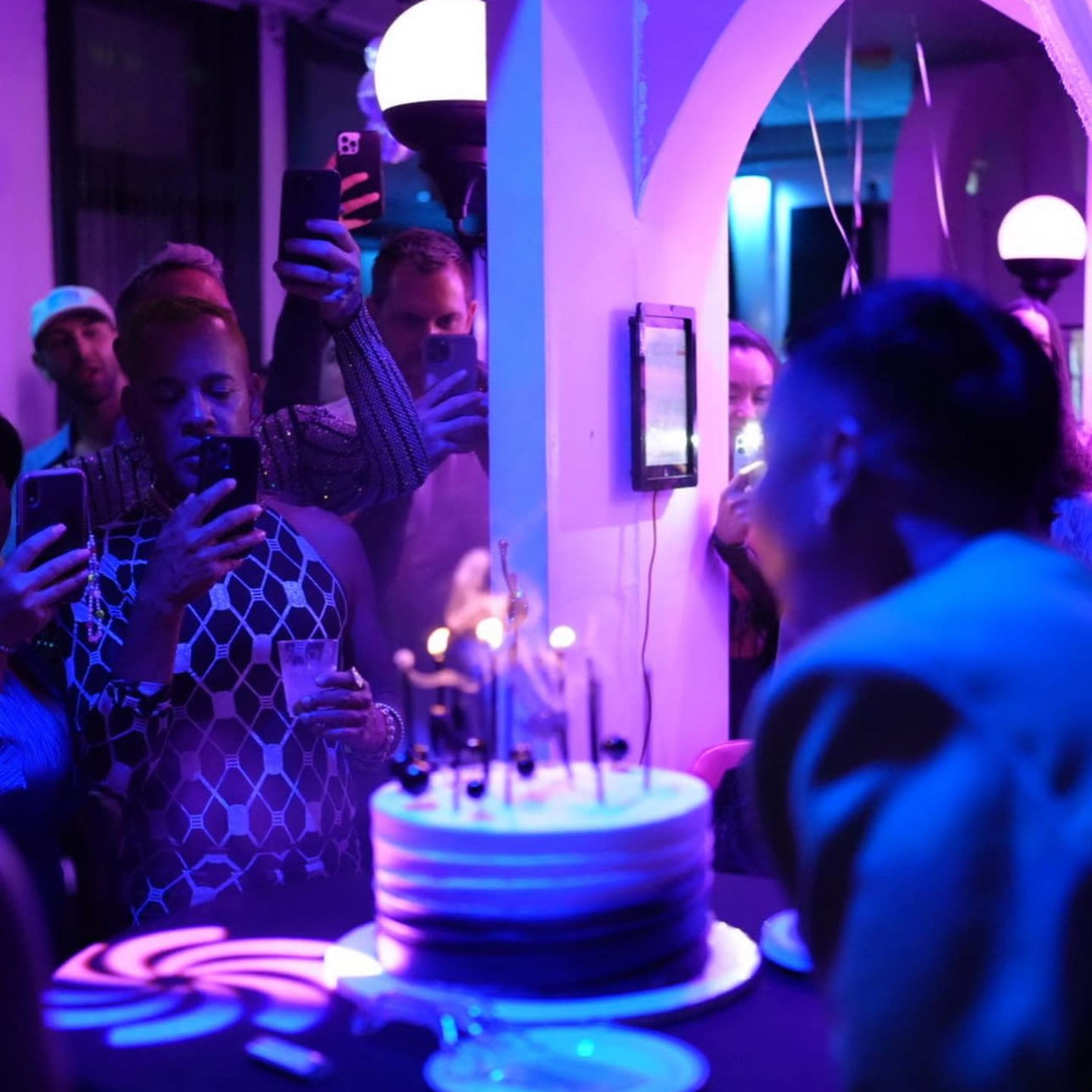 A group of people gathers around a table with a large cake adorned with candles, while several attendees capture the moment on their phones.