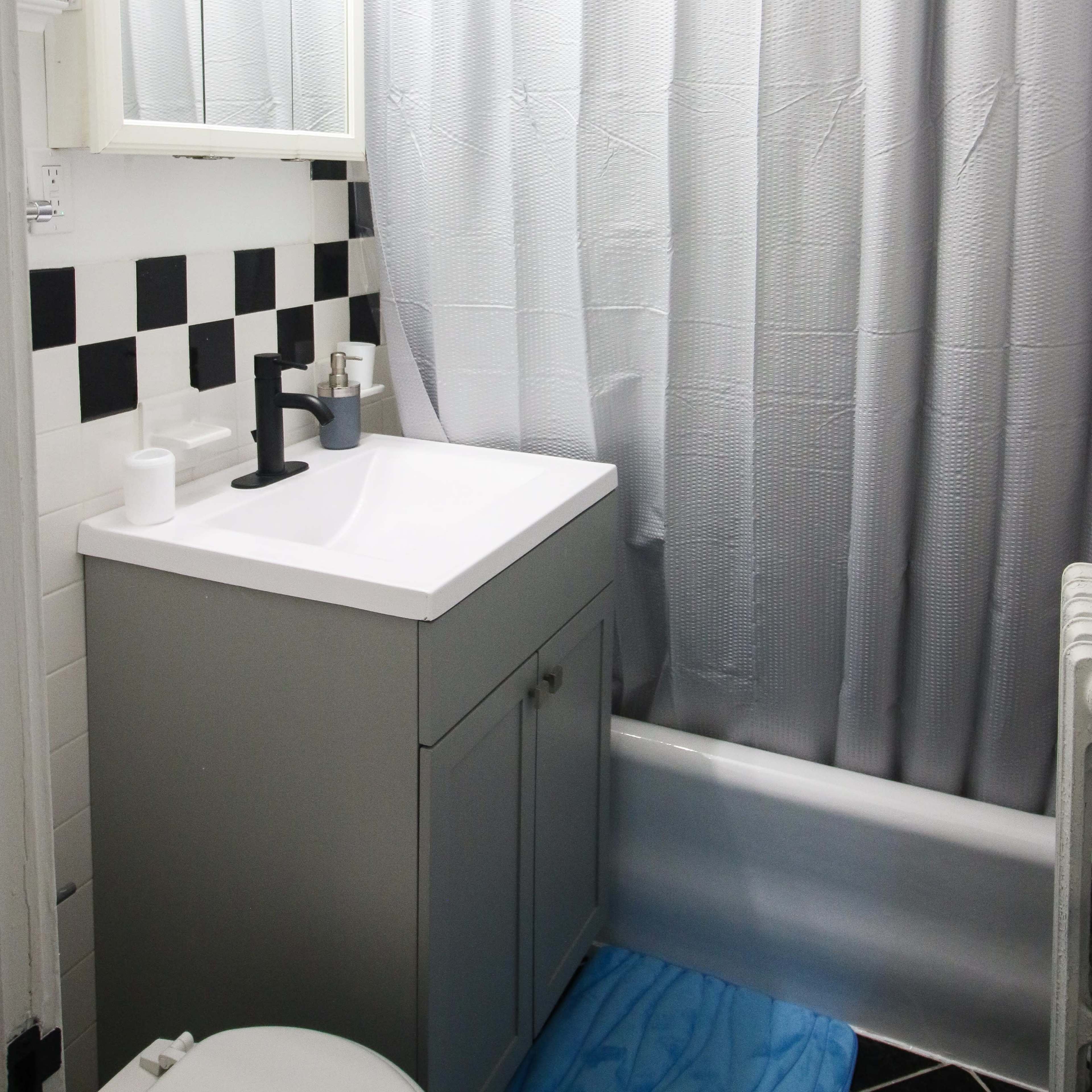 A bathroom with a gray vanity cabinet, a white sink, a white shower curtain, and a black and white tiled floor.