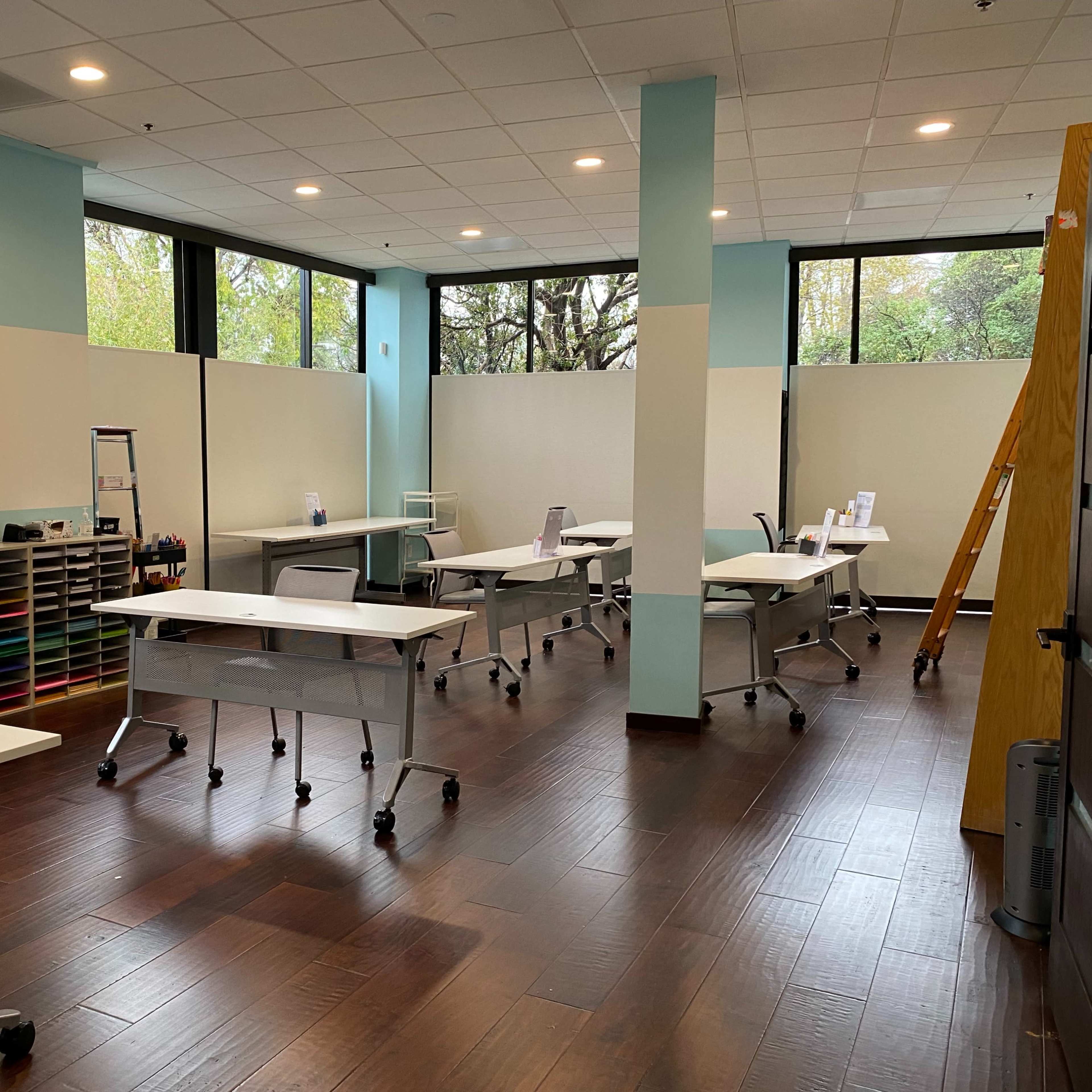 The room features several white desks arranged in a spacious area with large windows and wooden flooring.