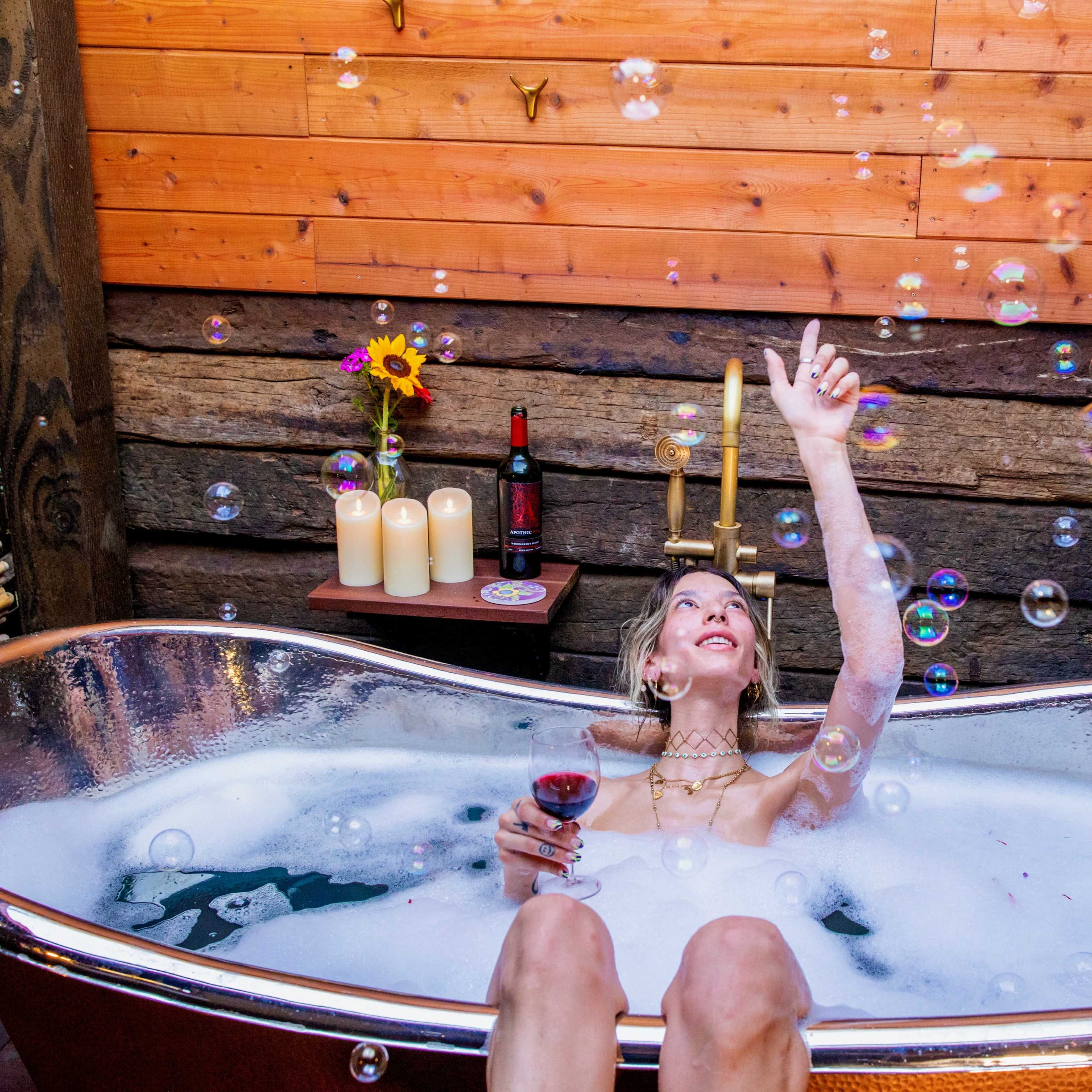 A person sits in a bubble-filled bathtub while holding a glass of wine and reaching for floating bubbles.
