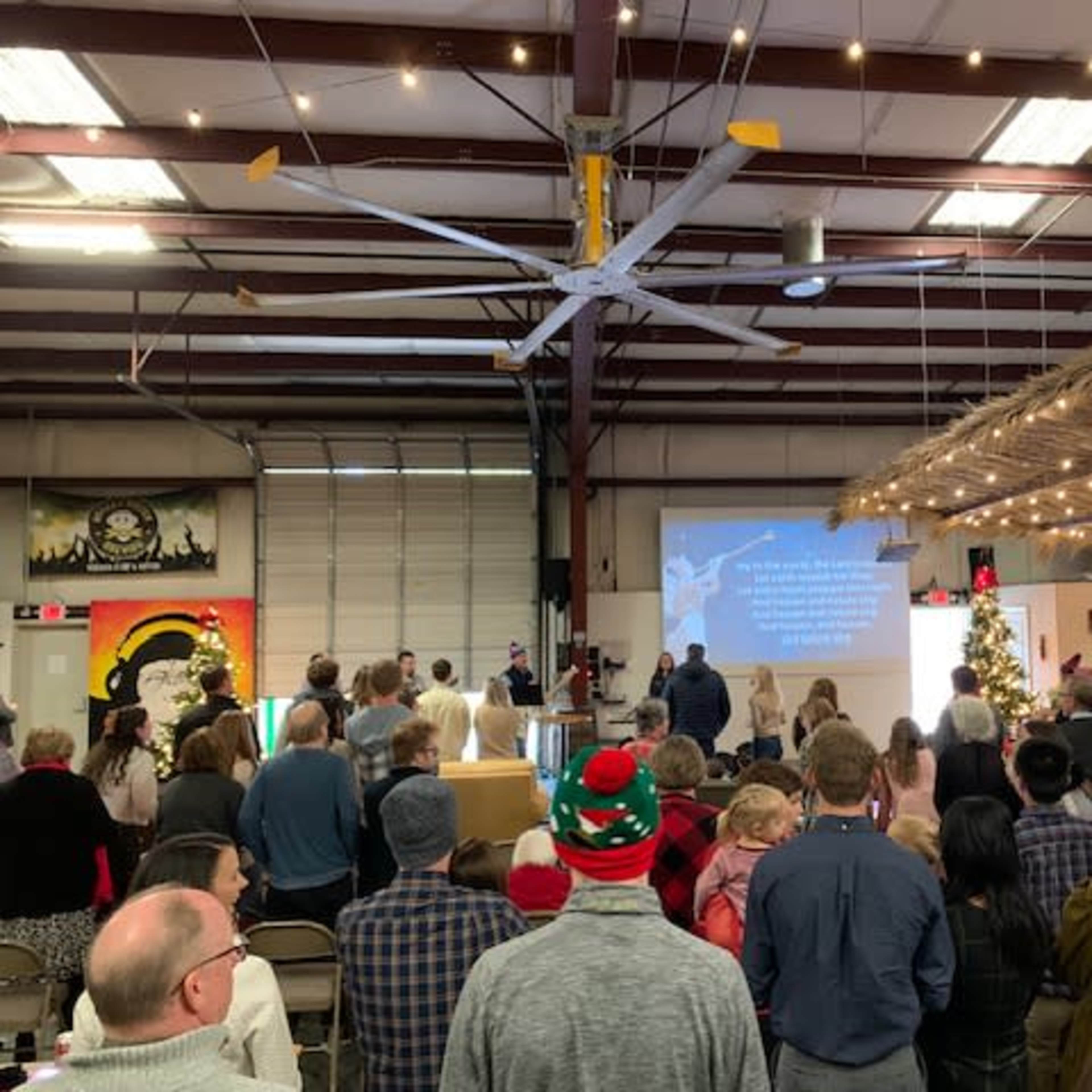 A large group of people stands in a warehouse-style space, facing a screen displaying text while festive decorations are visible around them.
