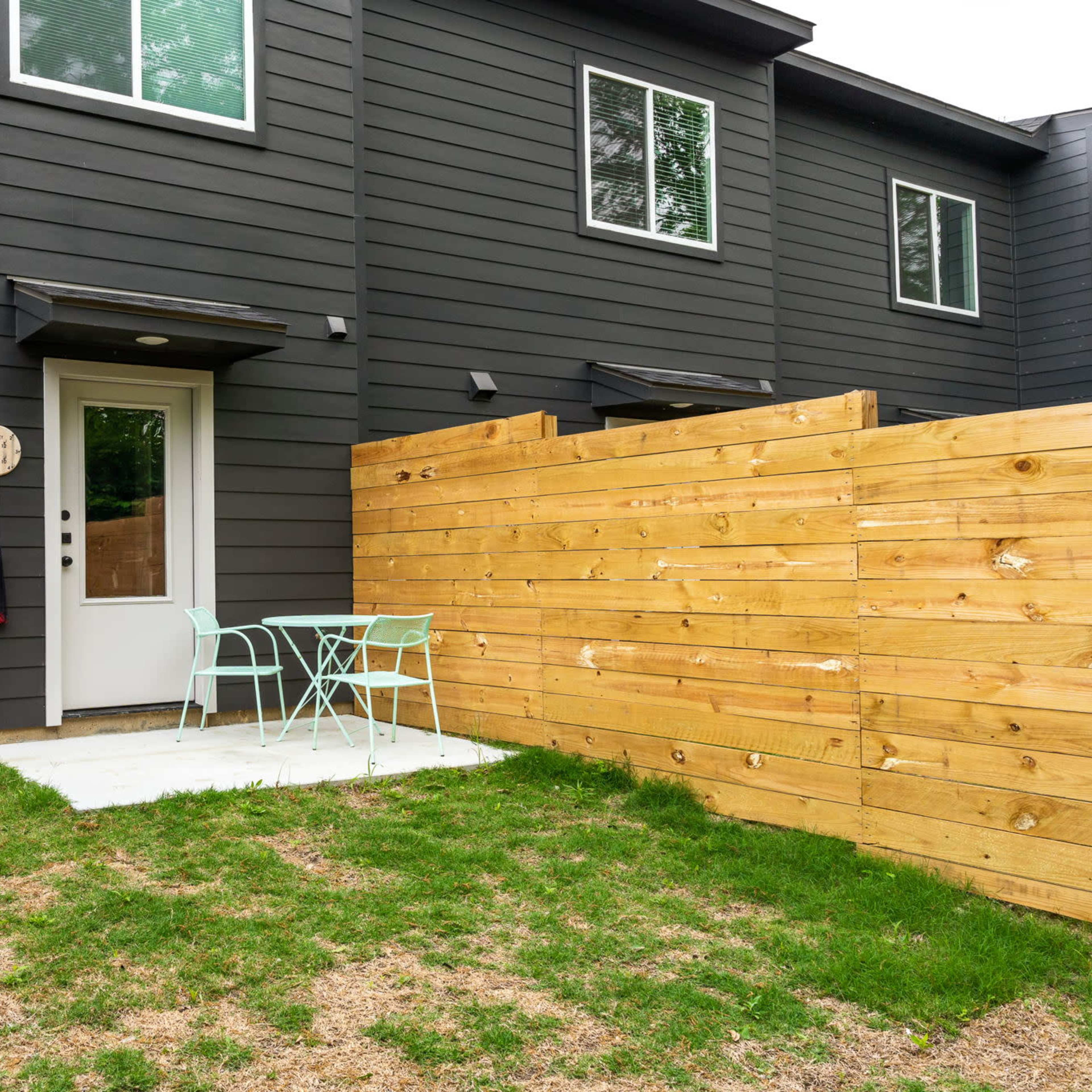 A small outdoor patio area with a light-colored table and two blue chairs surrounded by a wooden fence and grass.