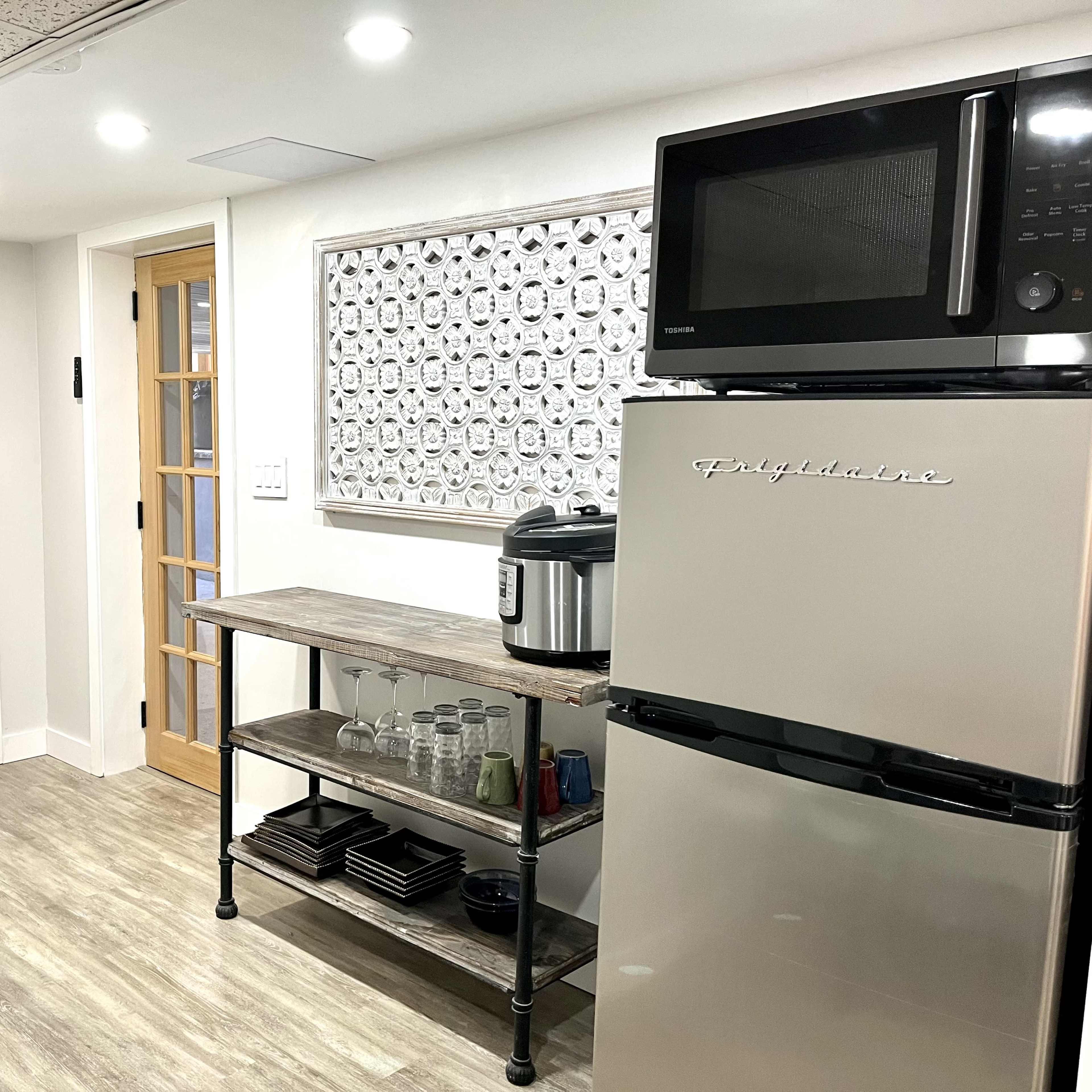 A modern kitchen with a stainless steel refrigerator, a microwave above it, a wooden shelf holding glassware and dishes, and a doorway leading to another room.