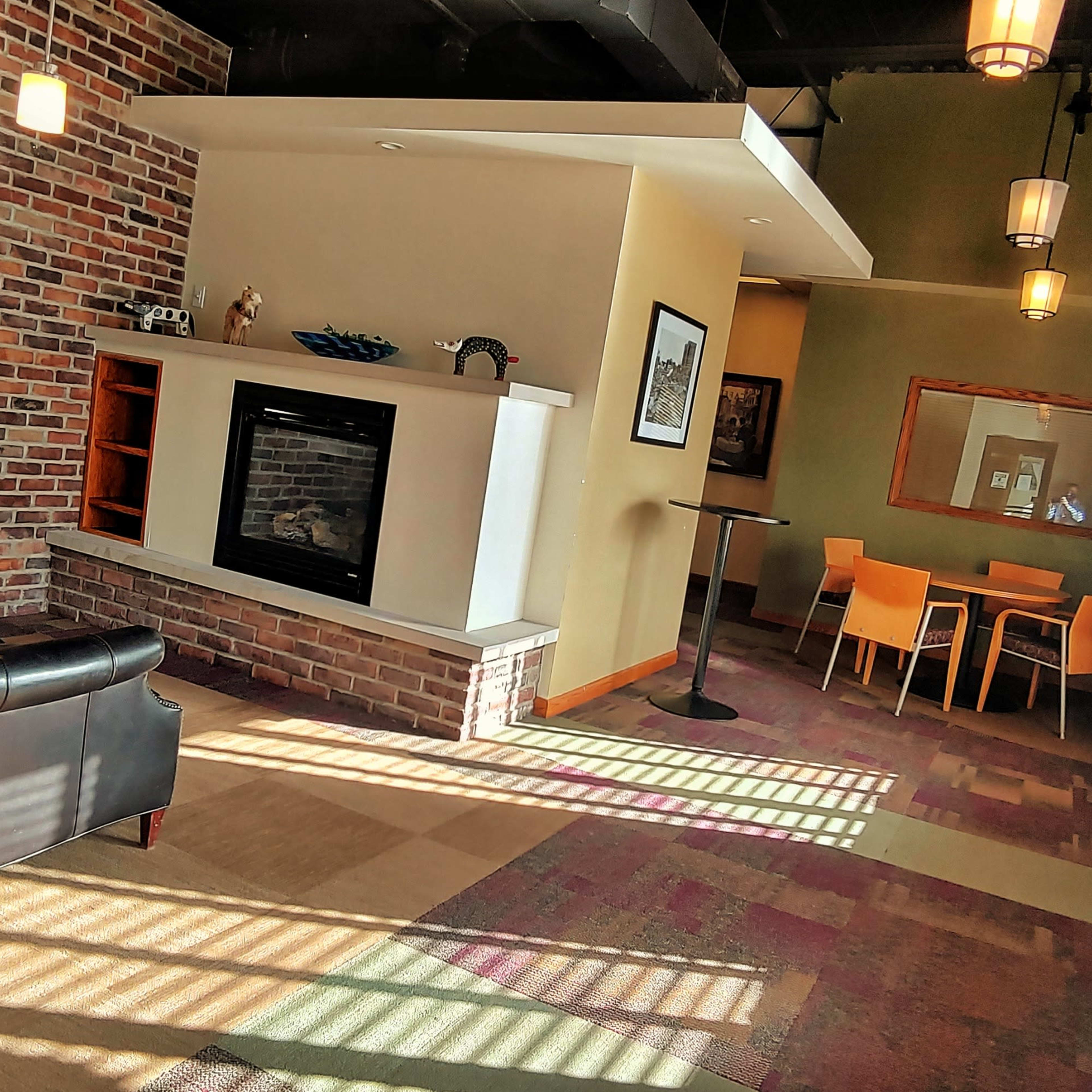 A cozy seating area with a black couch, a brick accent wall, a fireplace, and a dining table set against a backdrop of warm sunlight streaming through the windows.