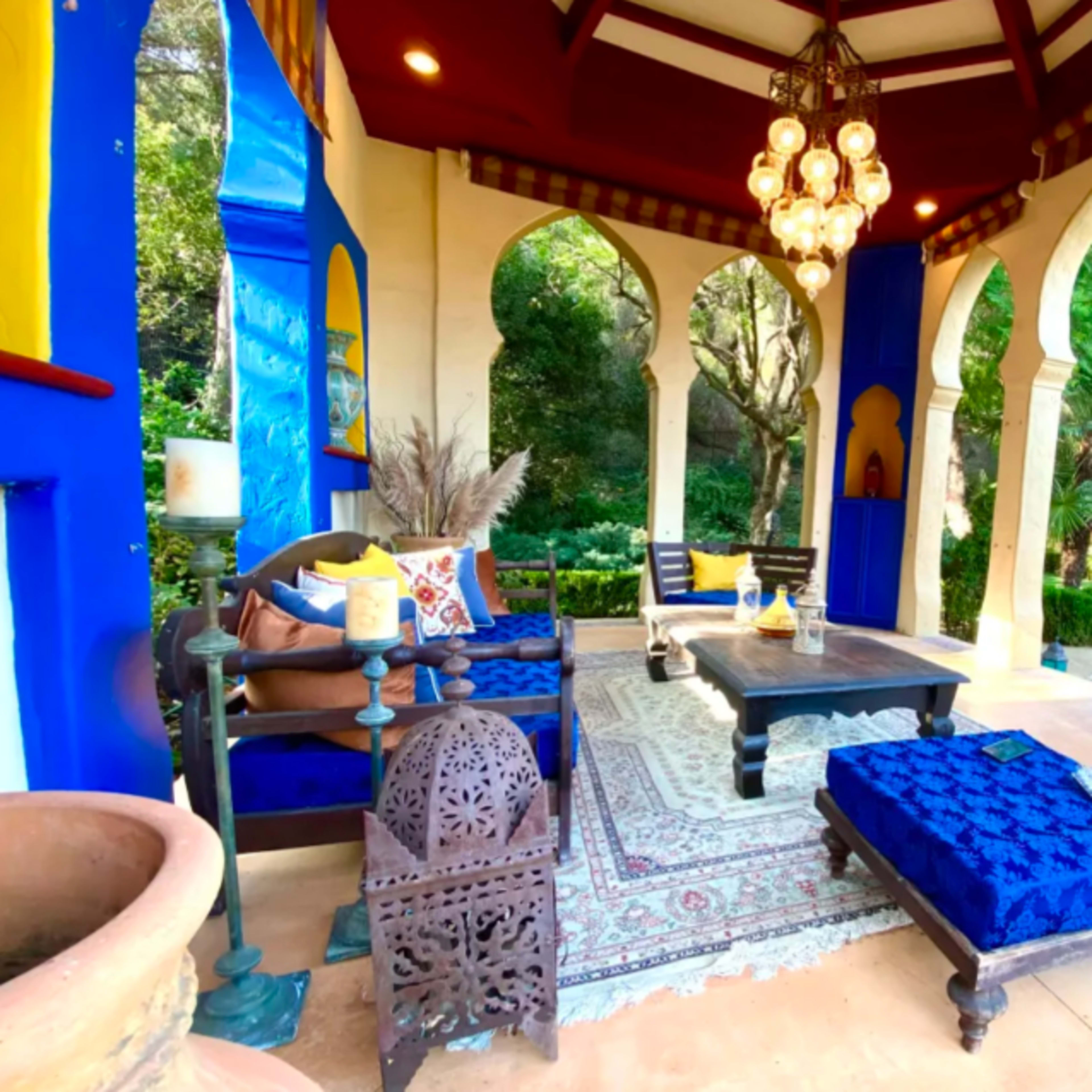 A brightly colored outdoor seating area with arches, featuring a dark wooden table, blue cushions, and decorative plants, set near a pool surrounded by greenery.