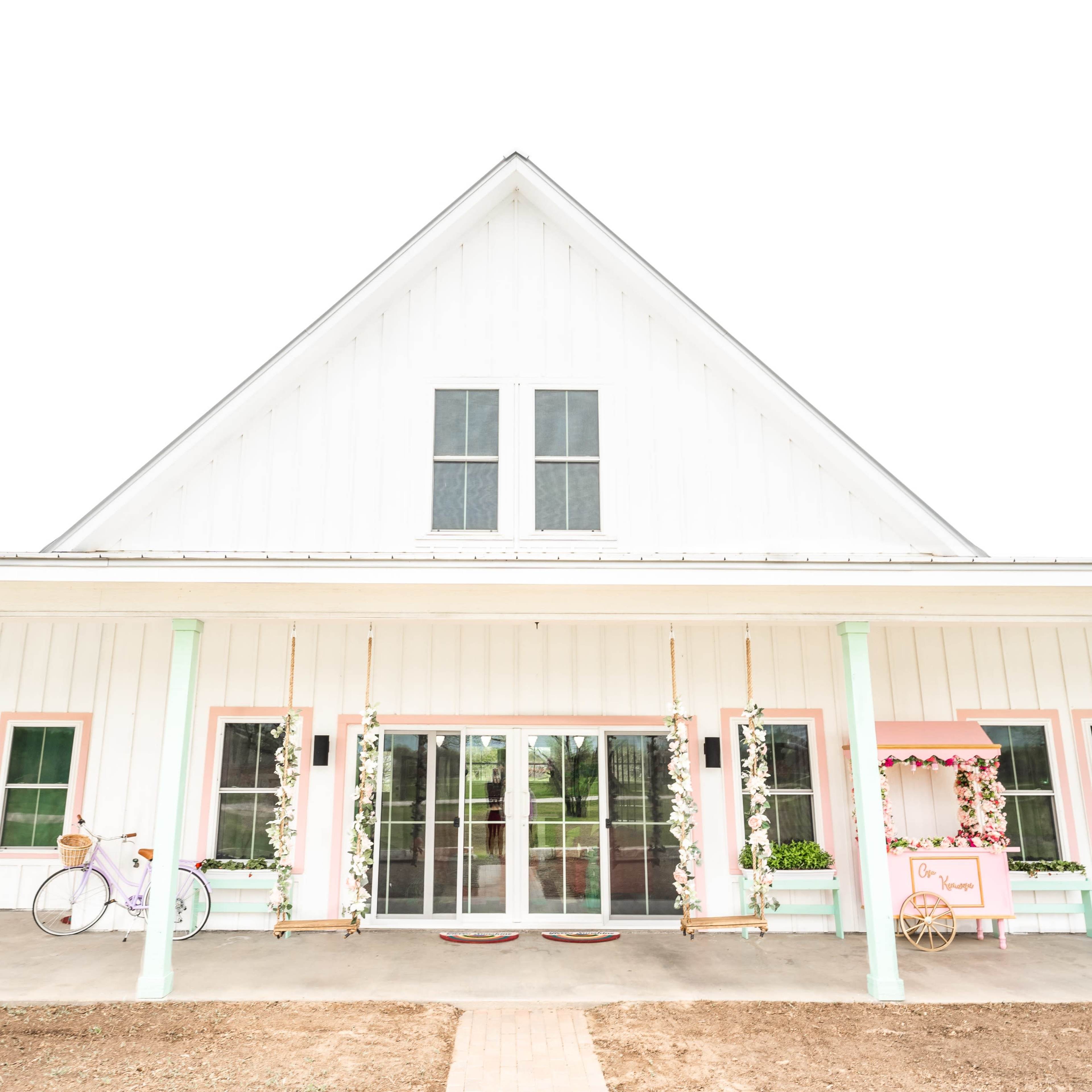 A white farmhouse with a peaked roof features large windows and a porch with swings and a decorative cart.