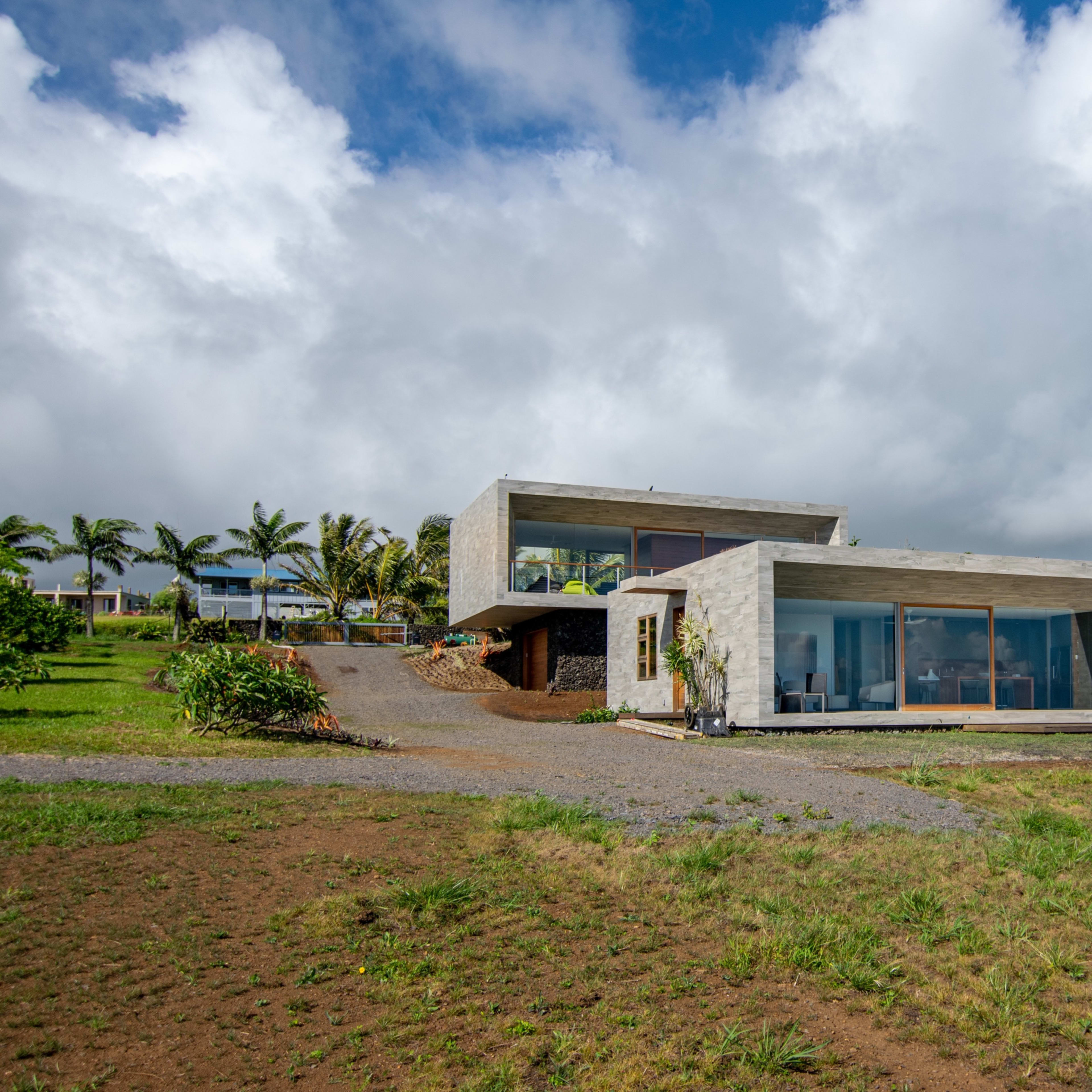 A modern concrete house with large windows is situated on a grassy landscape under a partly cloudy sky.