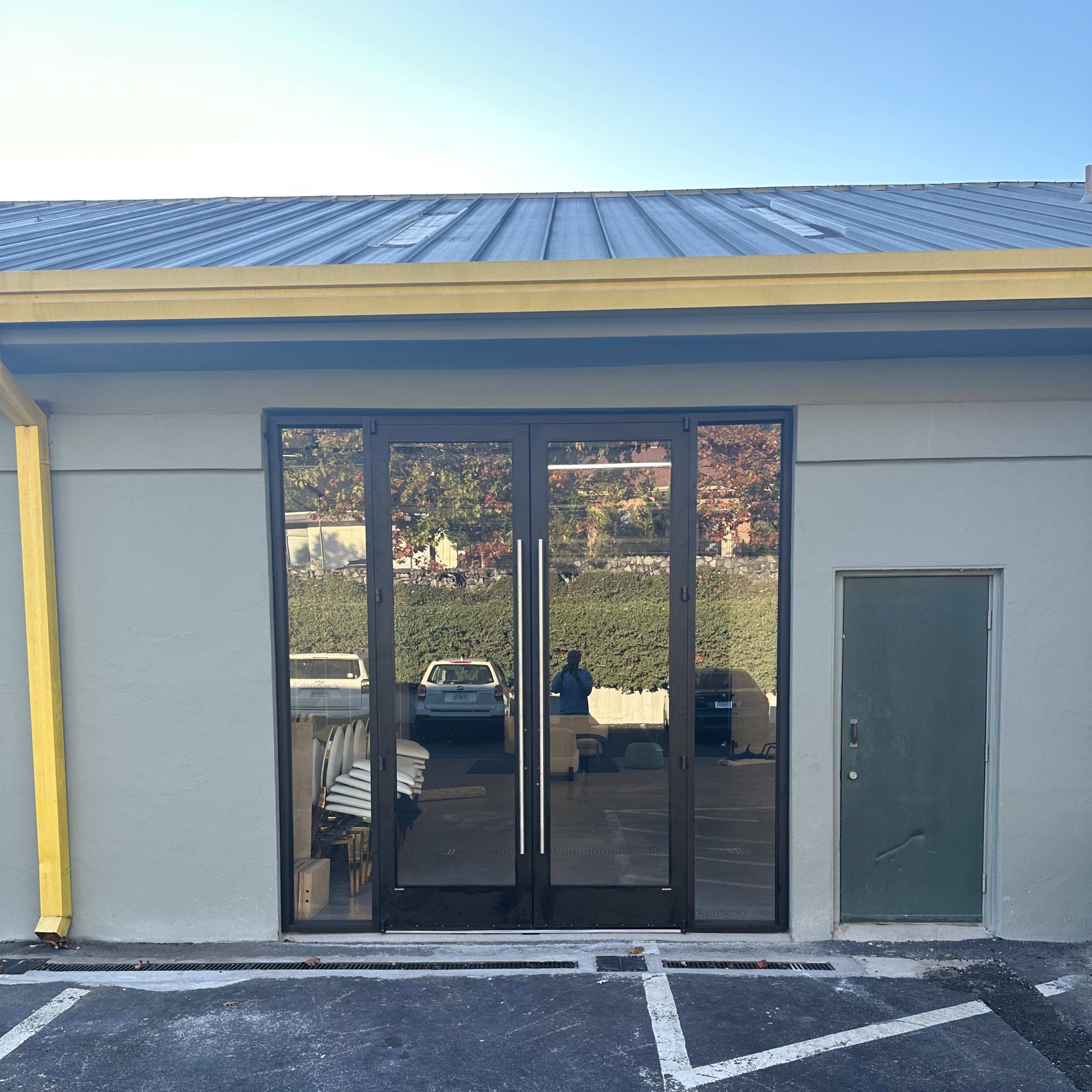 A building facade featuring large glass doors with a muted gray wall and a side door, all situated next to a parking lot.