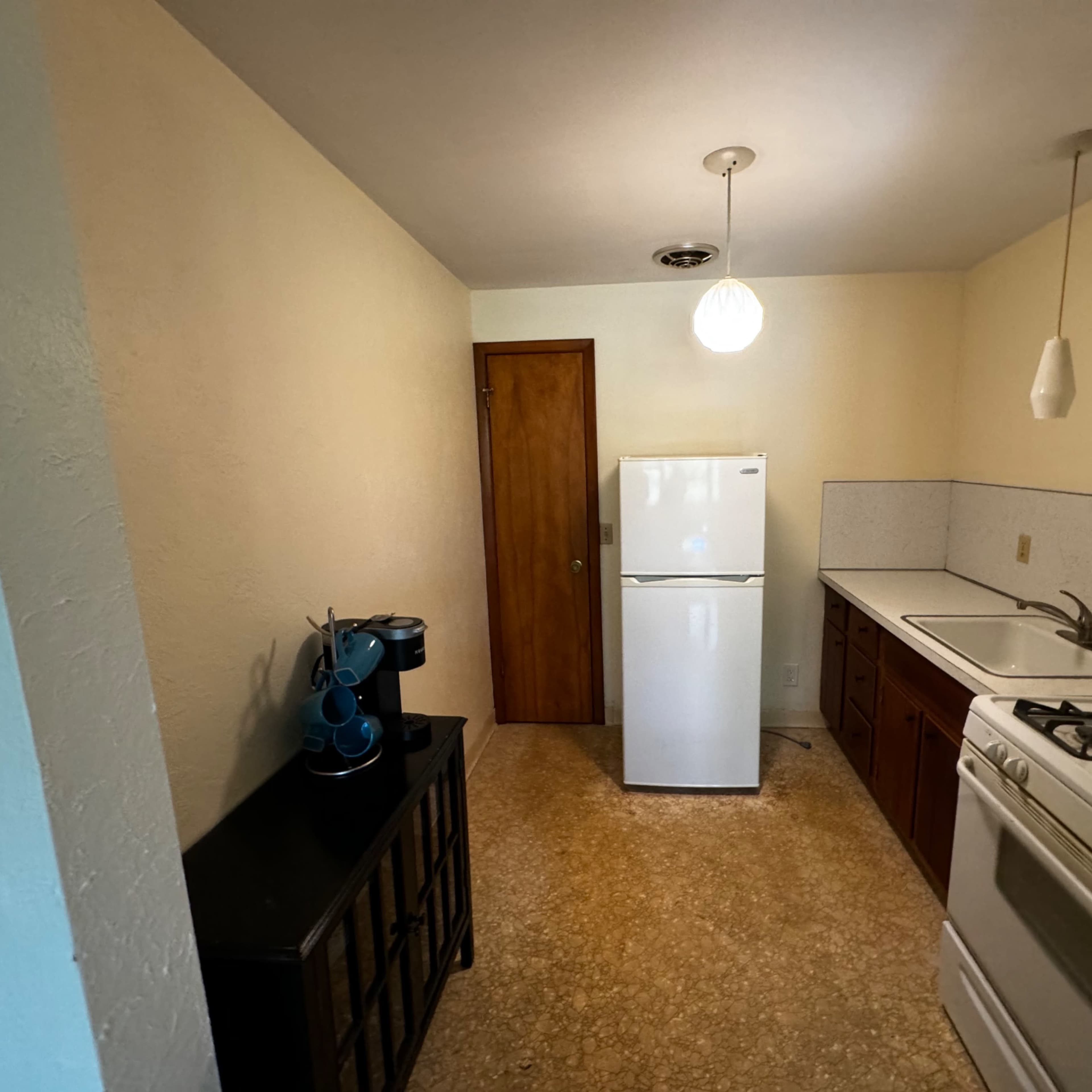 A small kitchen features a refrigerator, stove, and sink, with a doorway leading to another room and a pendant light hanging from the ceiling.