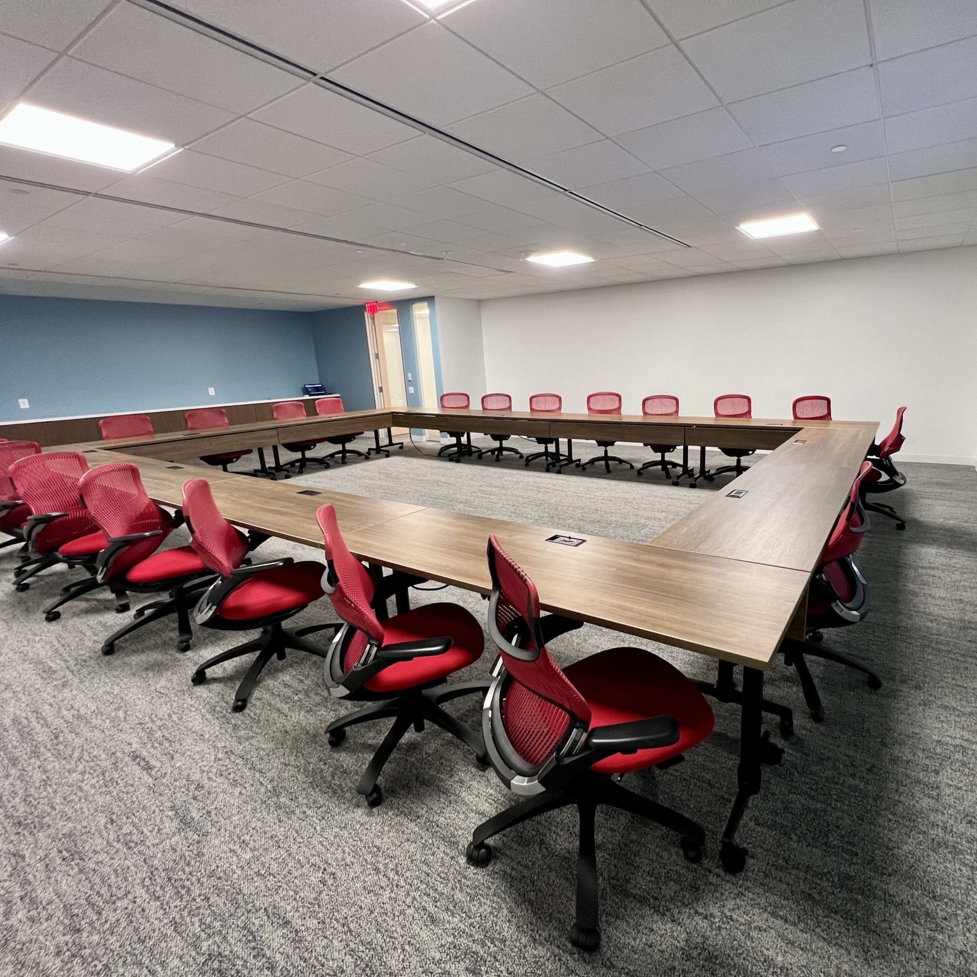 A large, rectangular conference table is arranged with red office chairs around it in a well-lit meeting room.