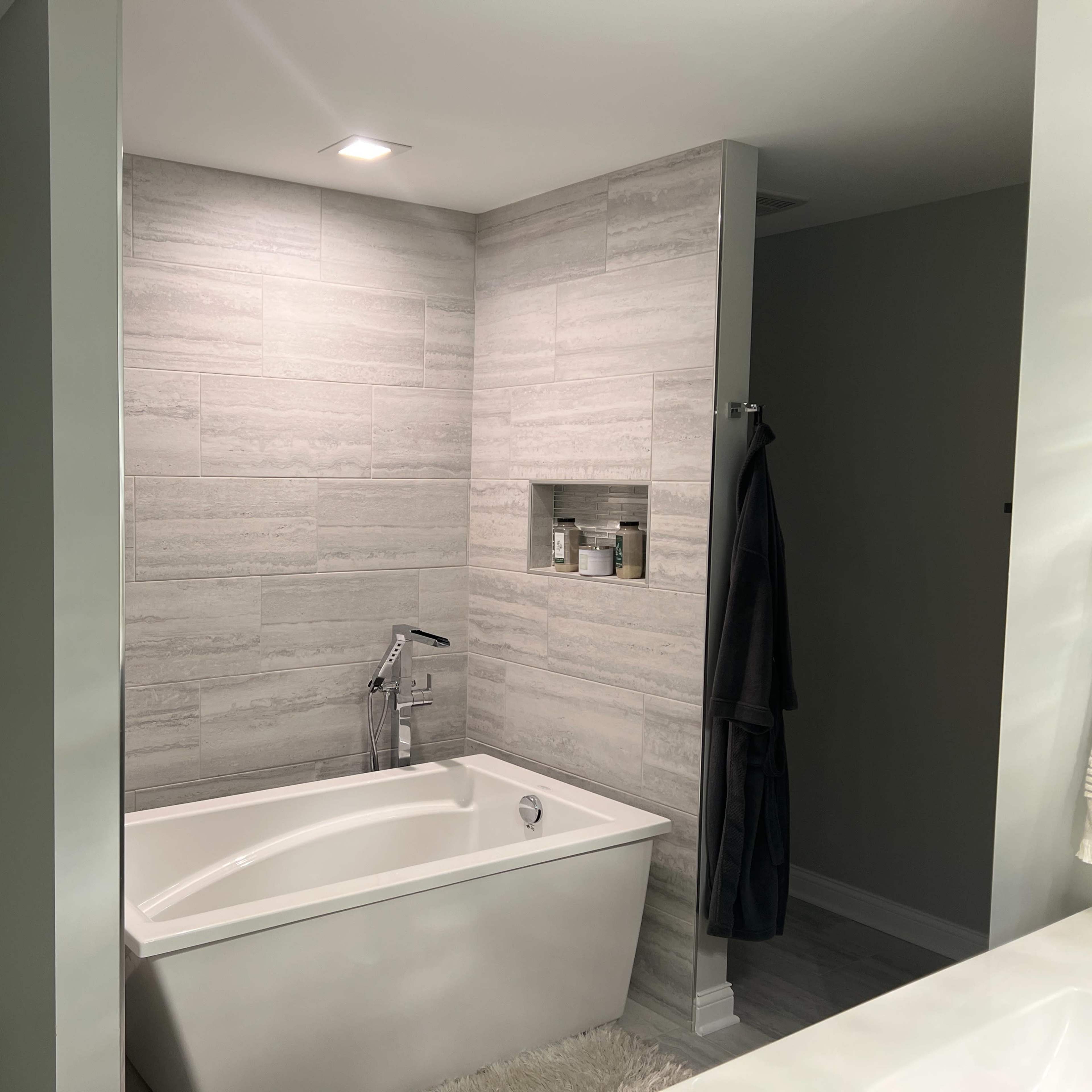 A modern bathroom featuring a freestanding bathtub, gray tiled walls, and a wall-mounted shelf with toiletries.