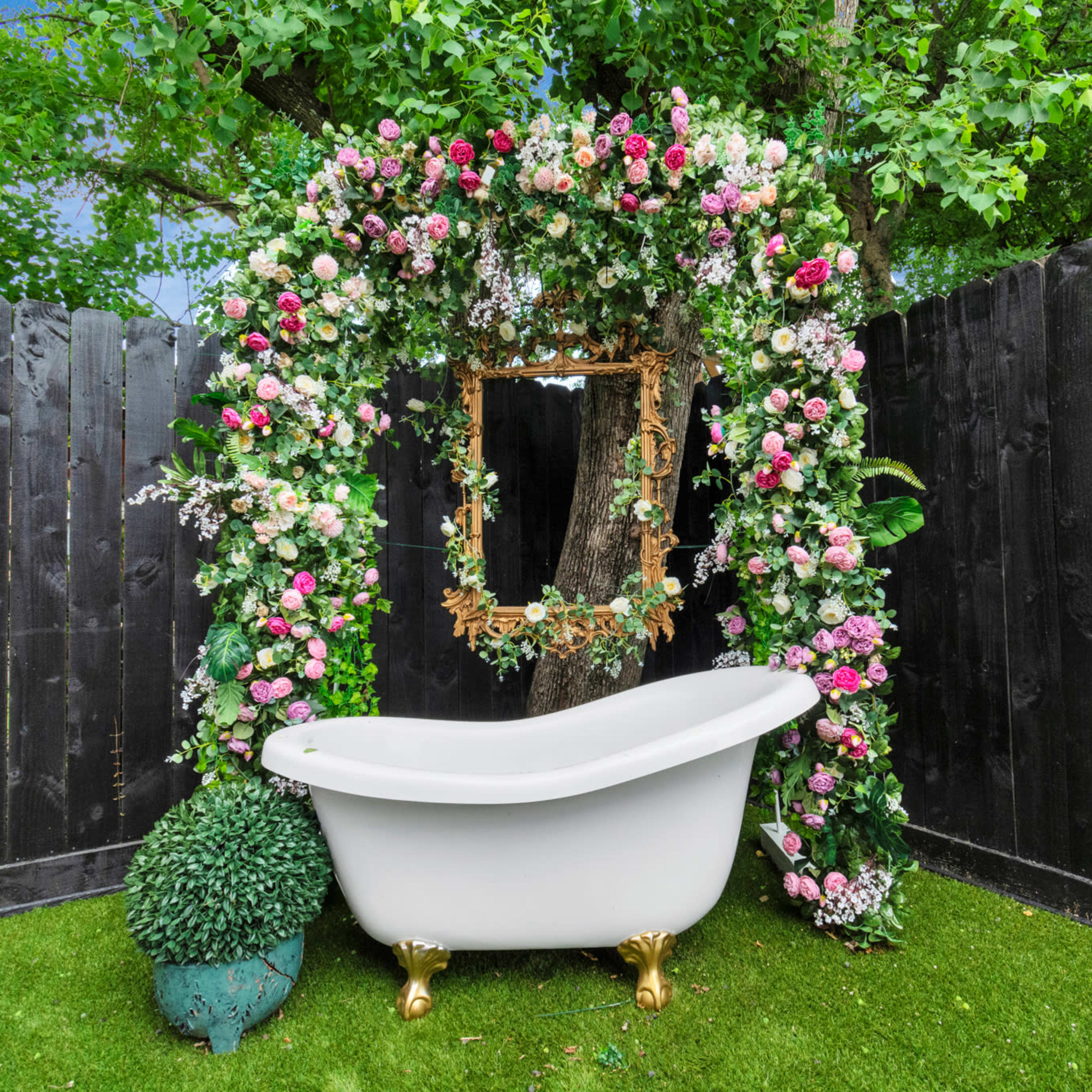 A white clawfoot bathtub is placed under a floral arch set against a backdrop of black wooden fencing and green foliage.