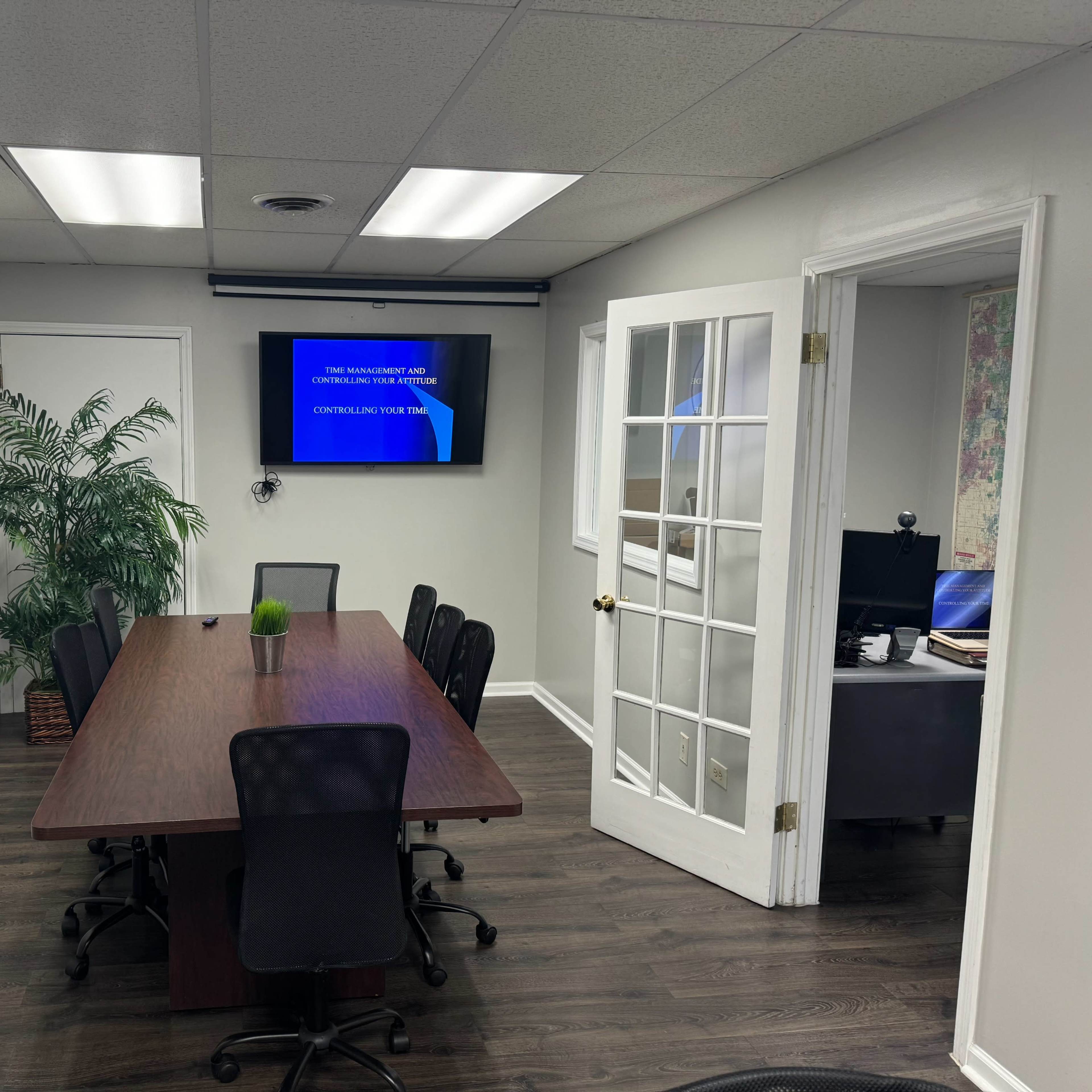 A conference room features a long wooden table surrounded by black chairs, with a television screen displaying text on one wall and an open door leading to another office.