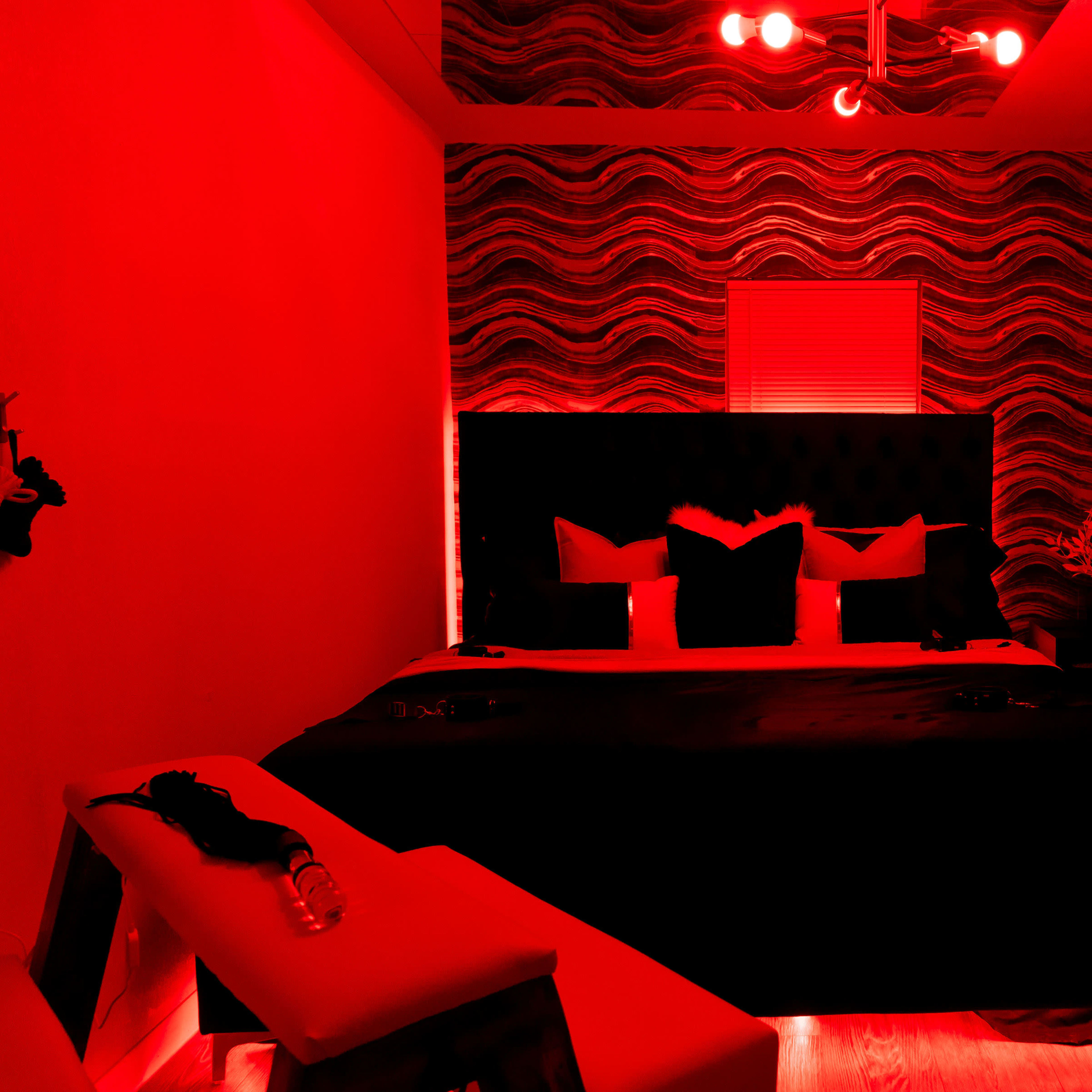A dimly lit bedroom featuring a large bed with dark bedding, decorative pillows, and a mannequin wearing chains, against a red-toned wall.