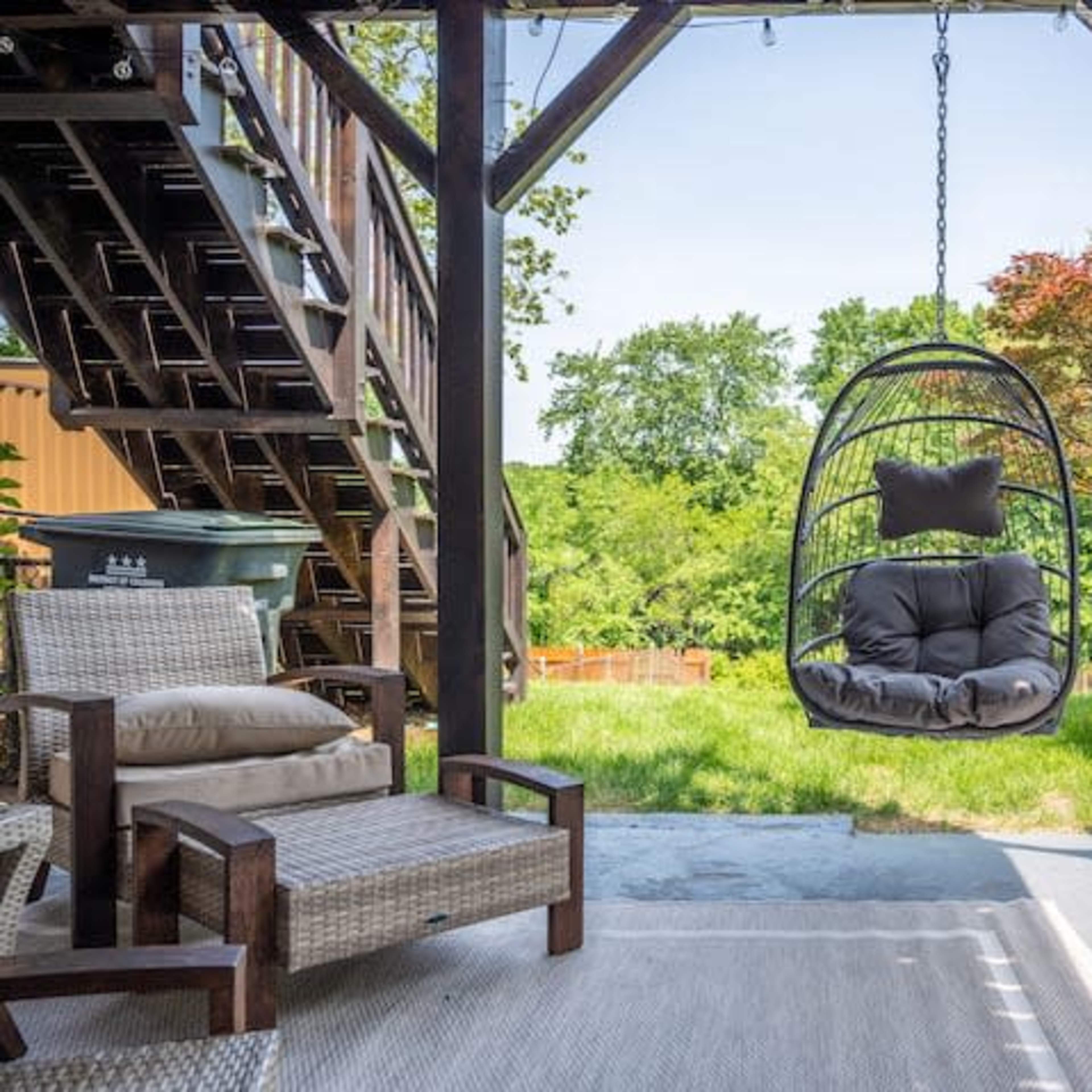 A hanging chair is suspended from a wooden structure in a backyard, surrounded by green trees and grass.