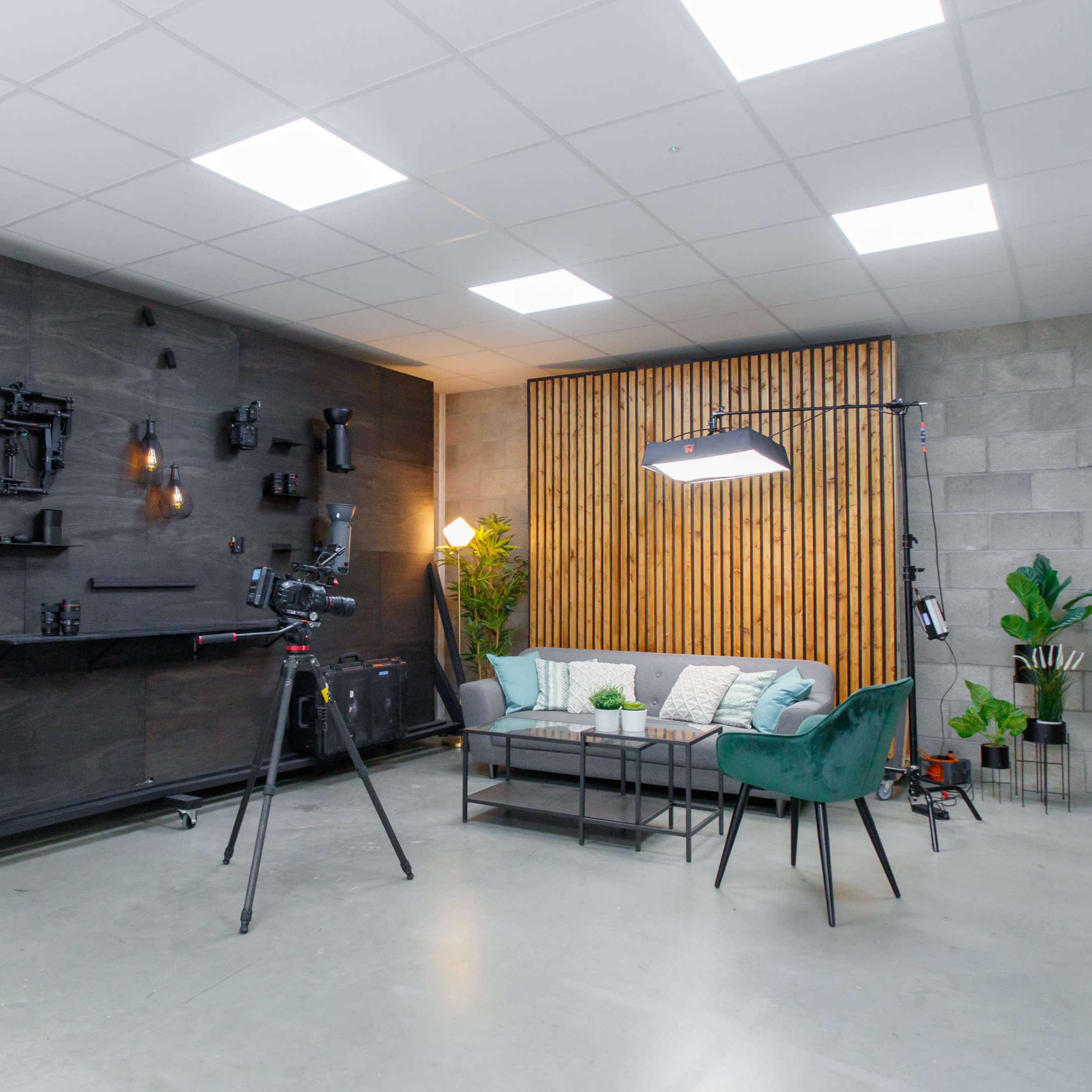 A modern studio setup with a couch, decorative plants, and a camera on a tripod against a backdrop of wooden slats and various equipment mounted on the wall.