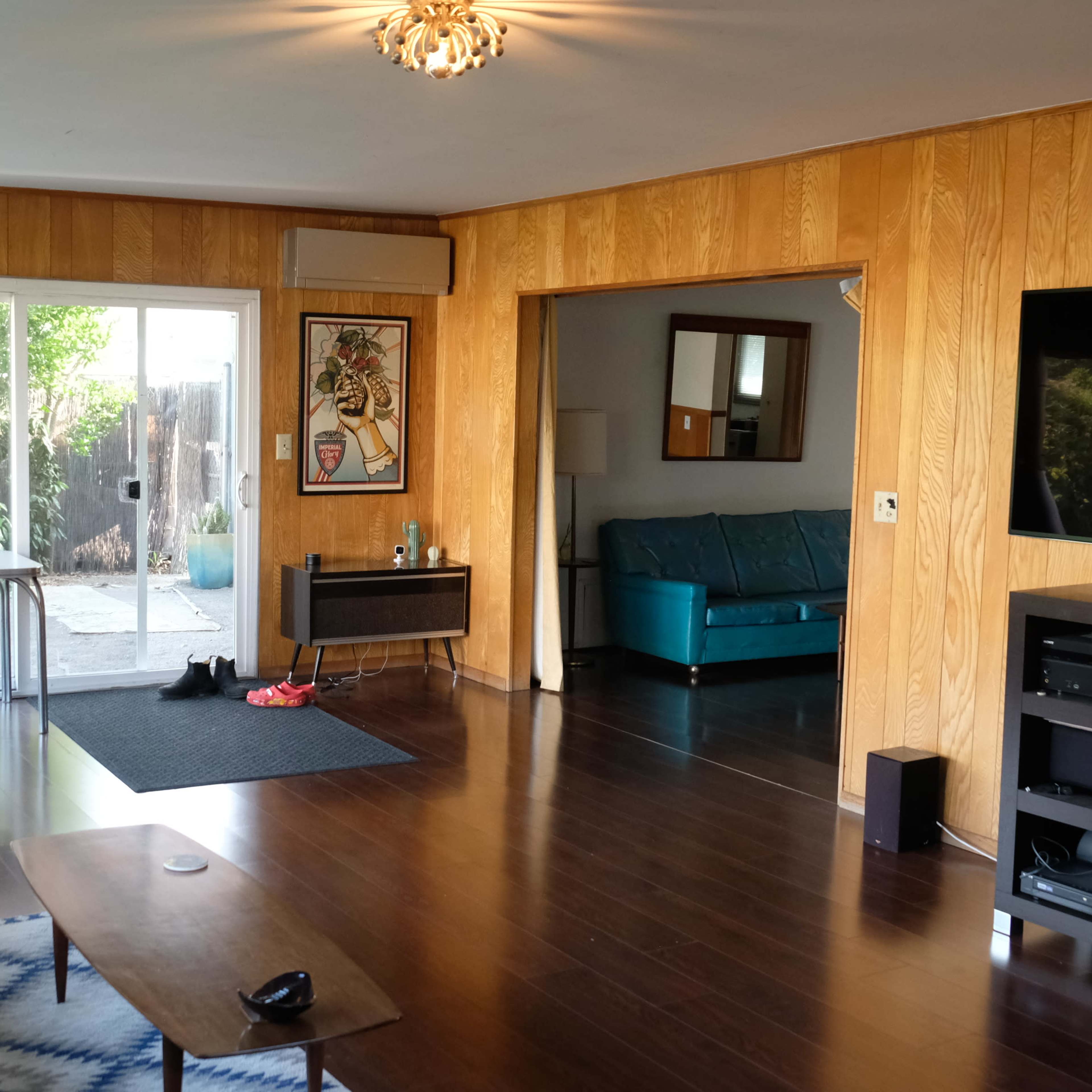A living room with wooden paneling, a large window facing a patio, and a flat-screen TV mounted on the wall opposite a couch and coffee table.