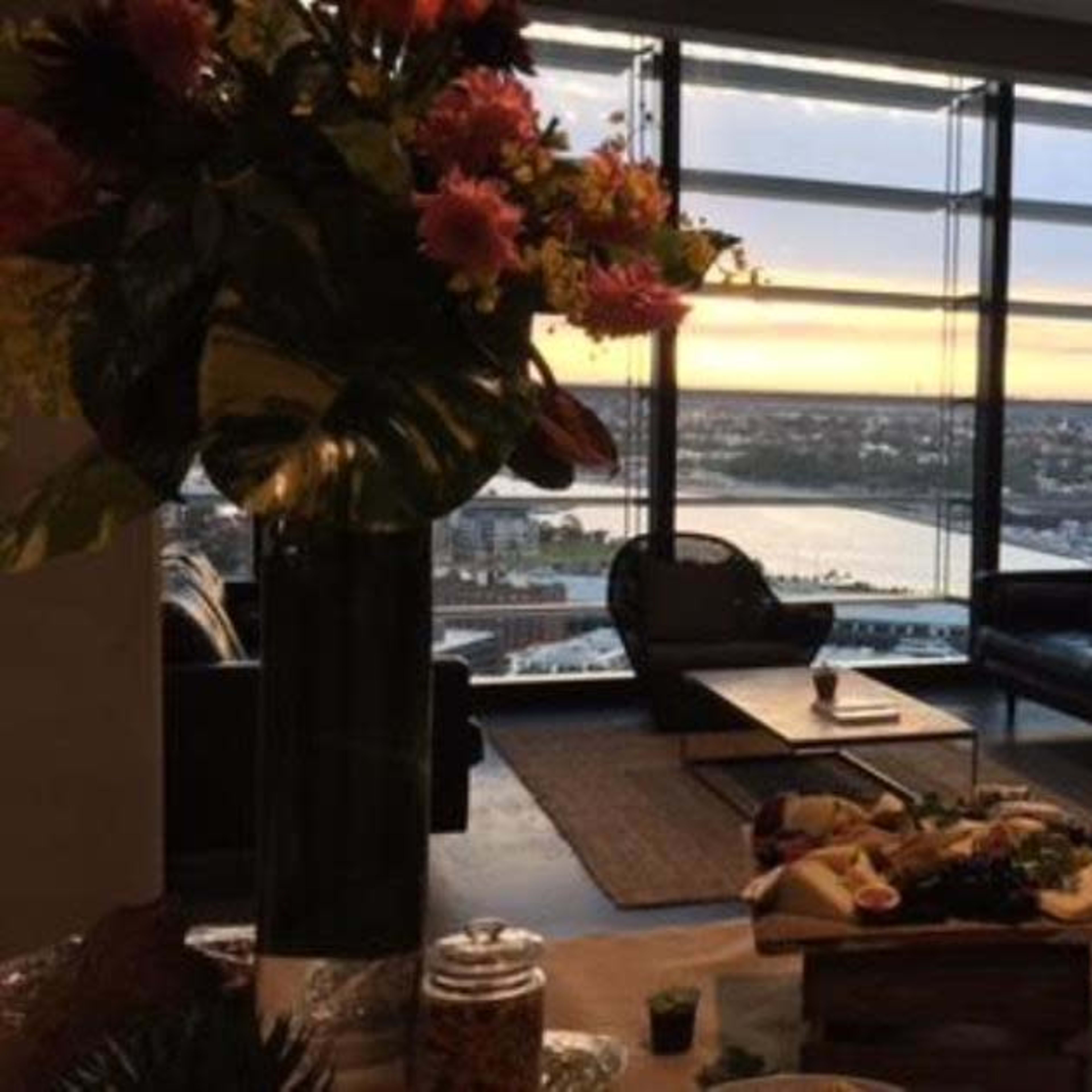 A modern interior with a buffet spread in the foreground, large windows displaying a sunset view over a body of water, and a figure standing by the window.