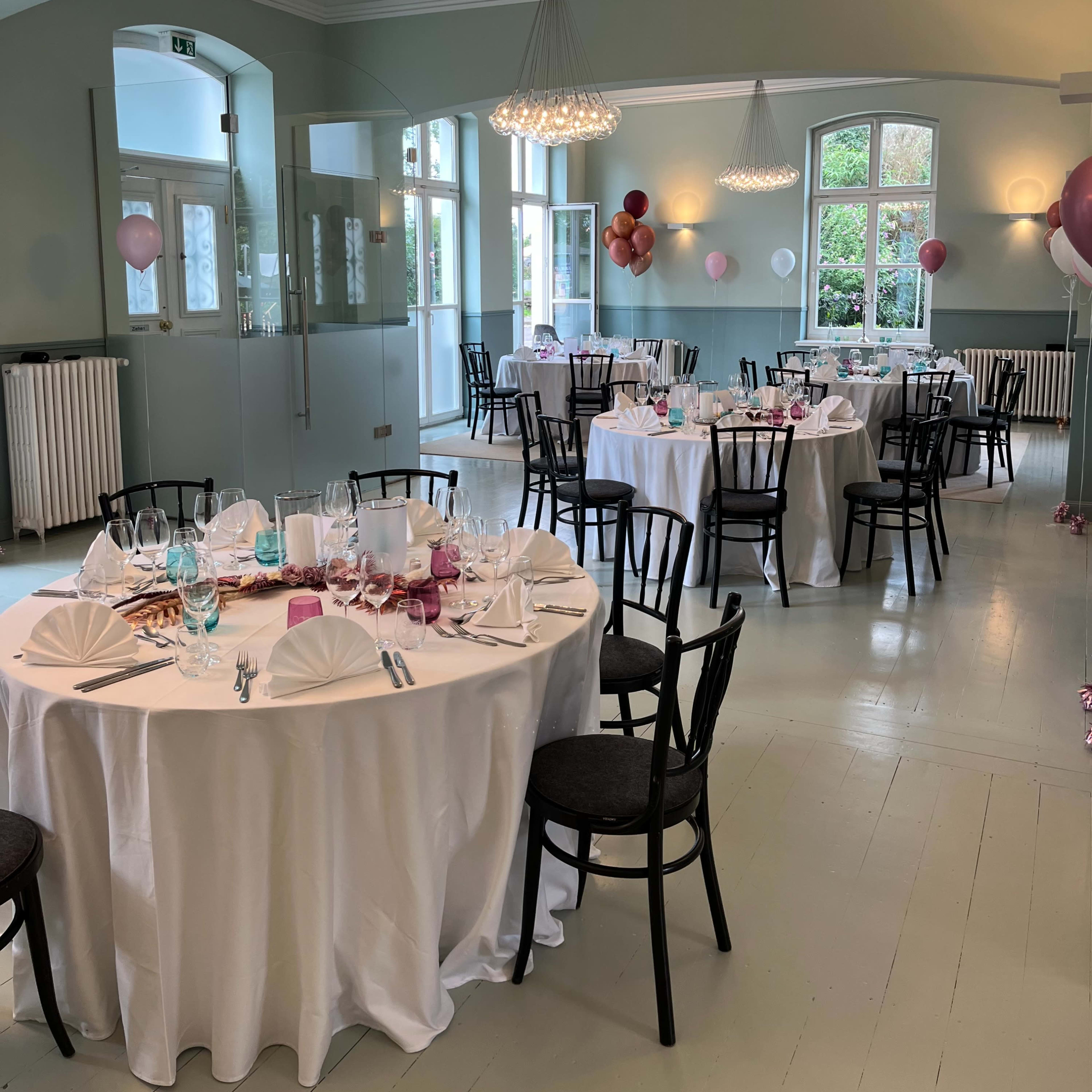 A banquet hall set for a dinner with round tables covered in white tablecloths and decorated with folded napkins, surrounded by balloons hung from the walls.