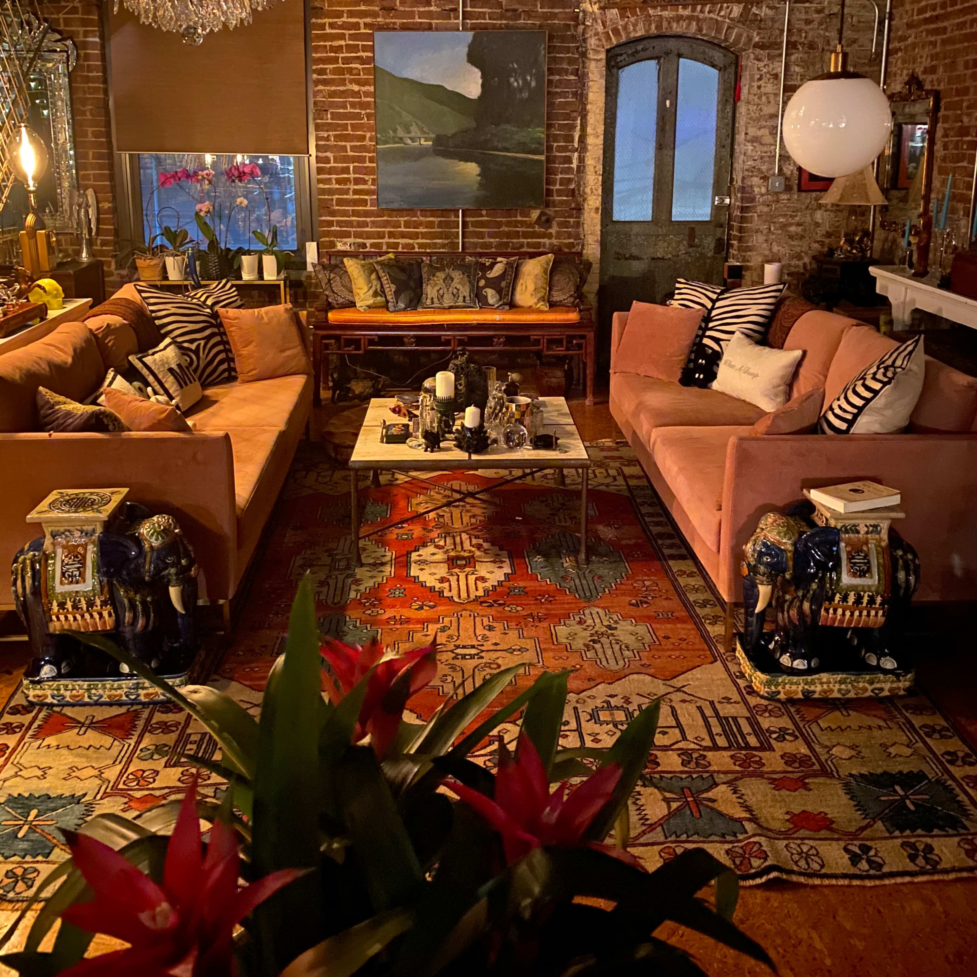 A cozy living room with two pink sofas facing each other on a richly patterned rug, surrounded by exposed brick walls and decorative elements.
