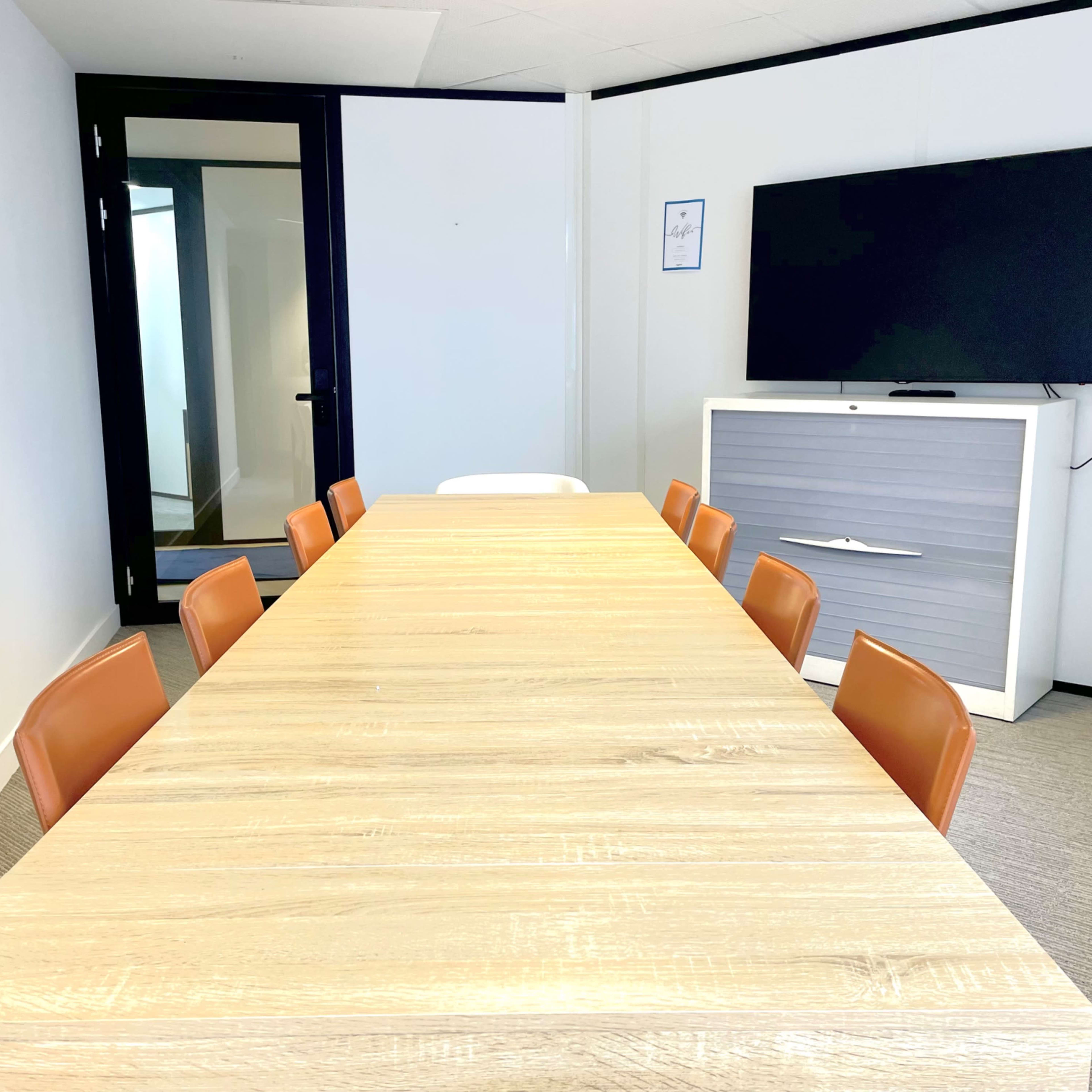 A long wooden conference table with eight orange chairs is positioned in a modern meeting room, facing a large wall-mounted television.