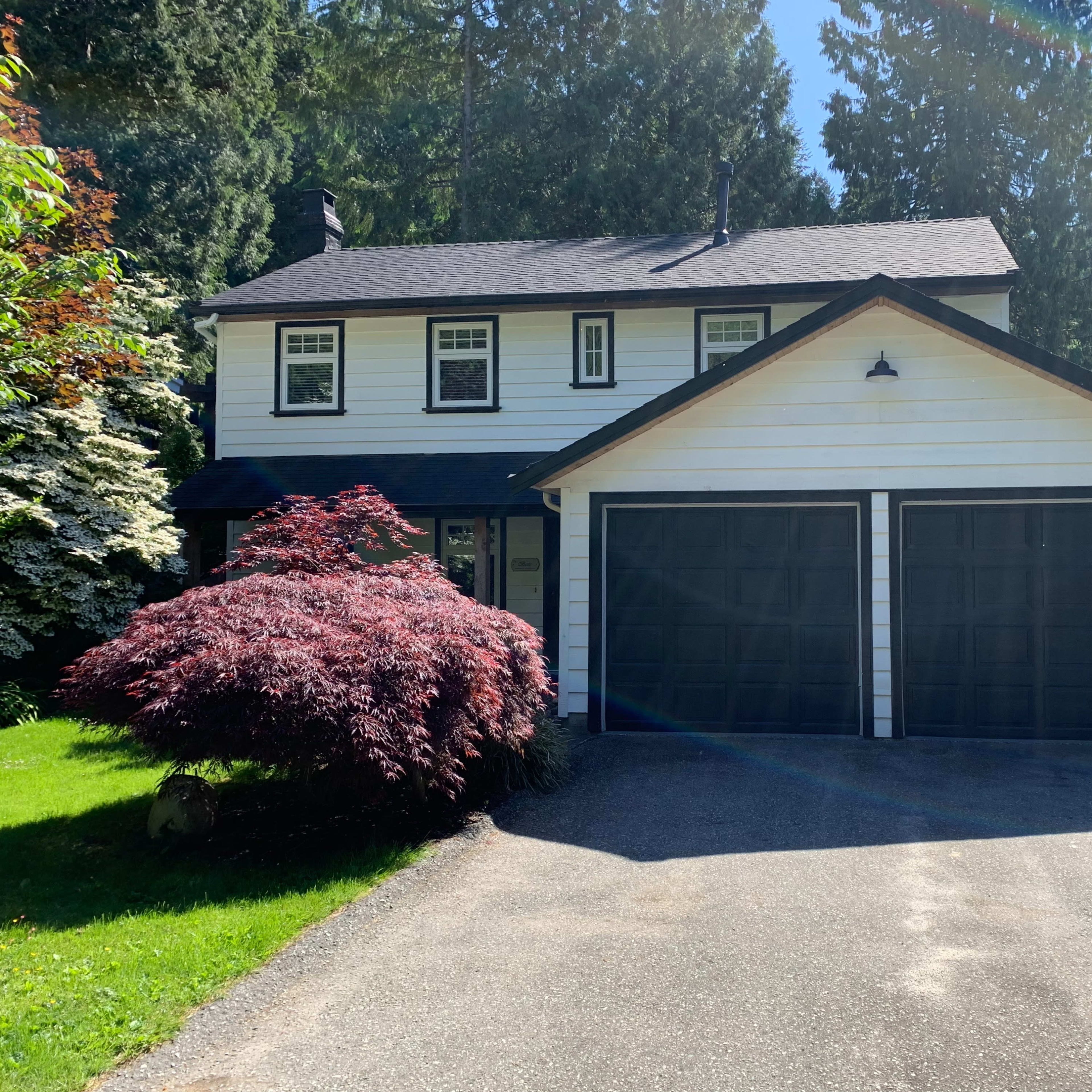 A two-story house with a black roof, white siding, and a double garage, surrounded by tall trees and shrubs.