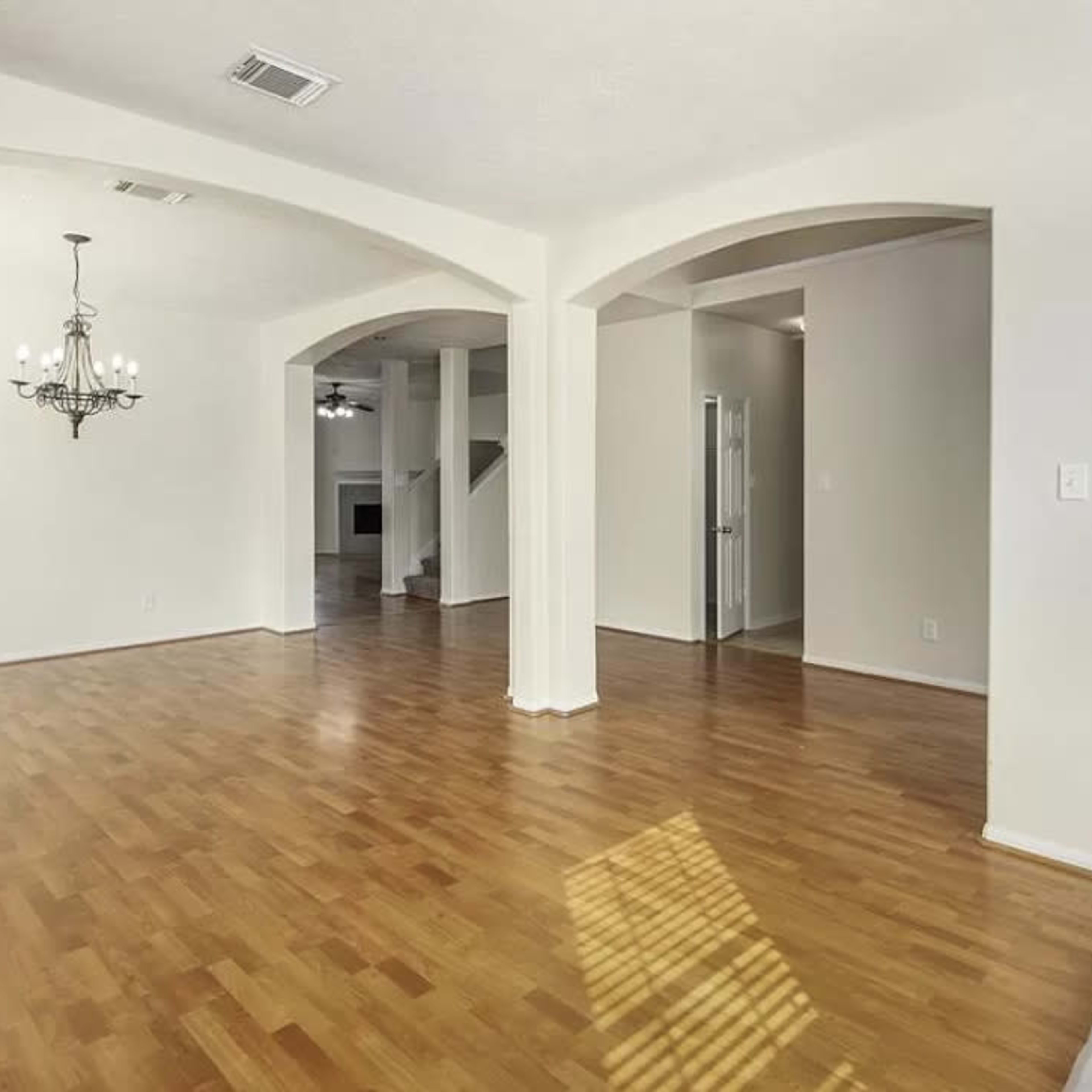 A spacious room with hardwood flooring, archway openings, and a chandelier, leading to a hallway and another area in the background.